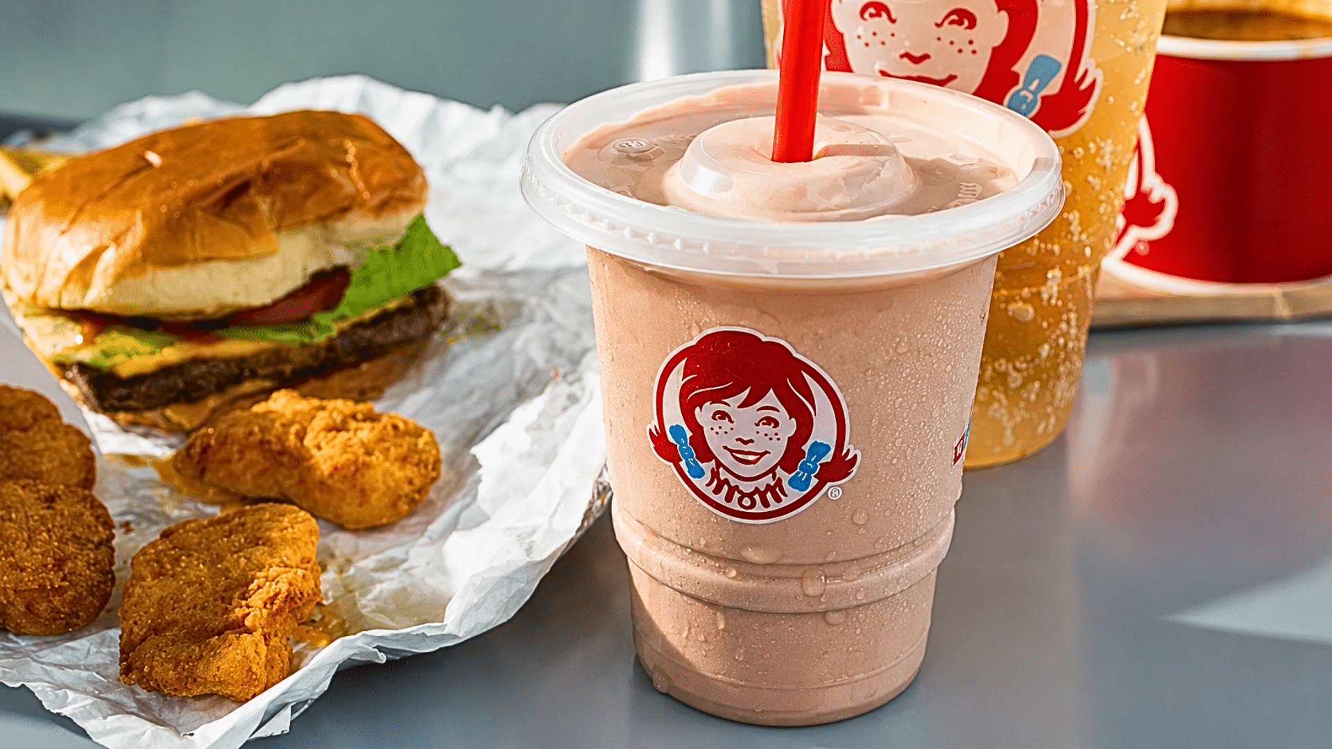 A close-up of a table spread featuring a large Chocolate Frosty in a clear plastic cup with a red straw; next to it is a freshly unwrapped cheeseburger with lettuce and tomato, four crispy chicken nuggets, and a fountain drink with the Wendy's girl logo visible in the background.