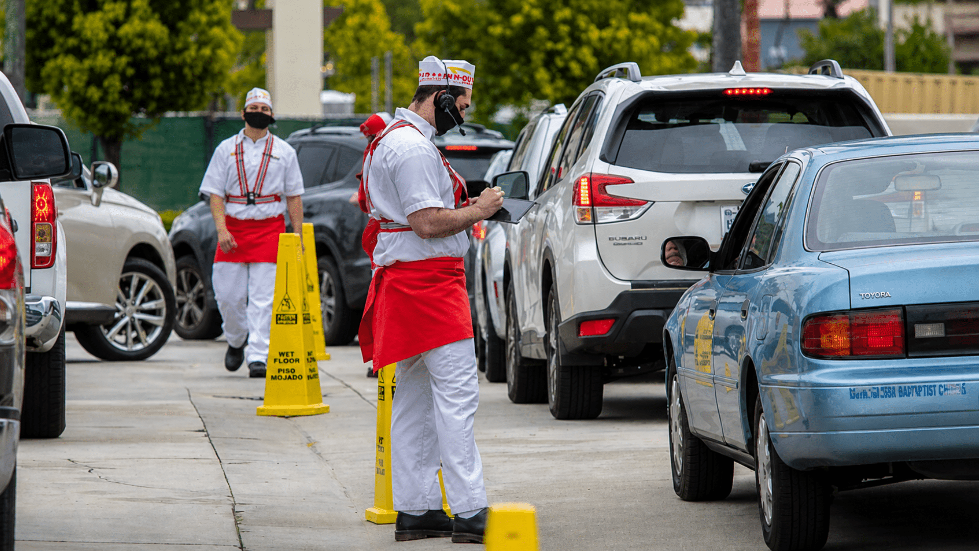 Two In-N-Out Burger employees in their signature white-and-red uniforms and paper hats are working a drive-thru line.