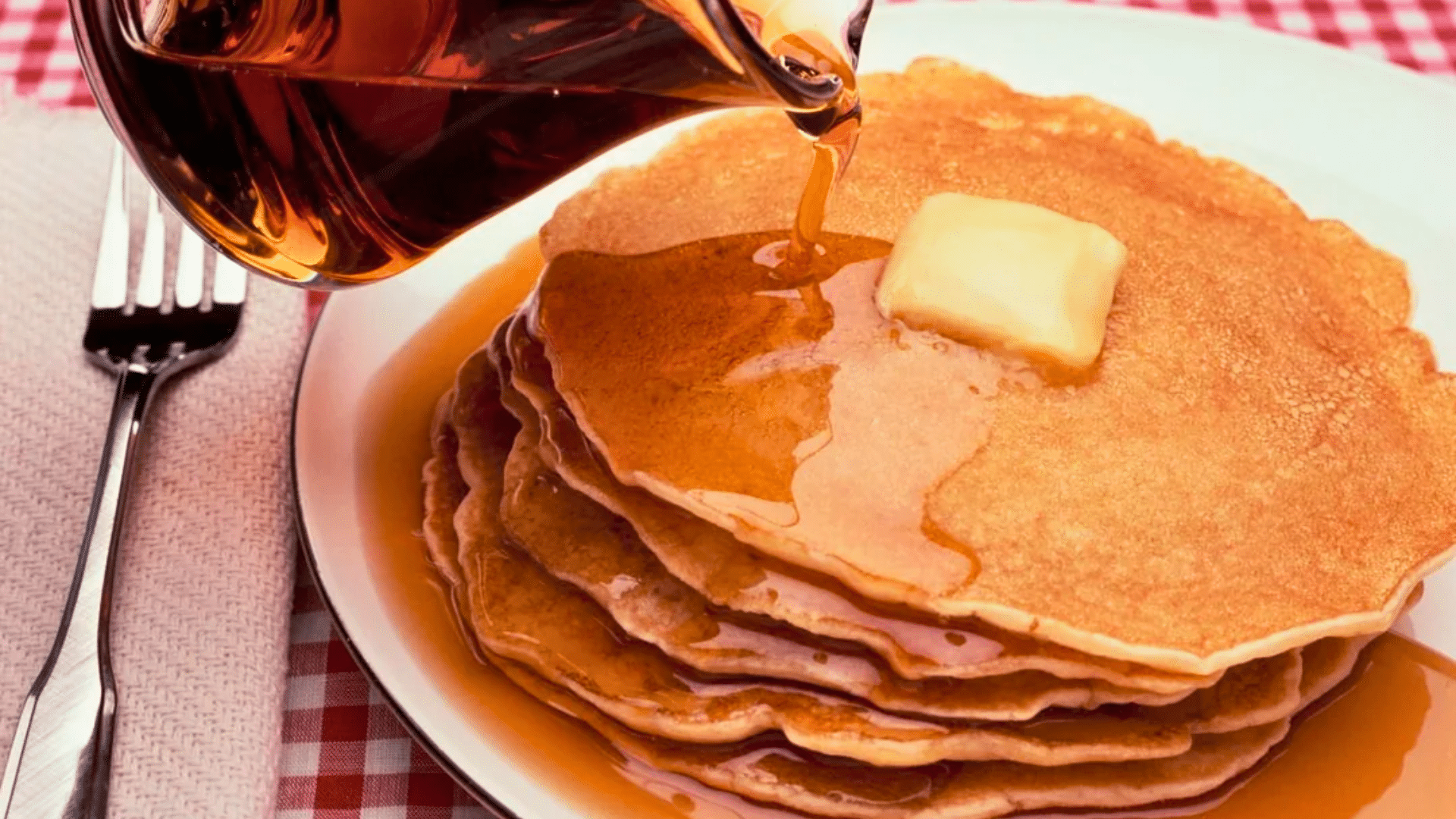 Syrup being poured over a stack of pancakes topped with butter.