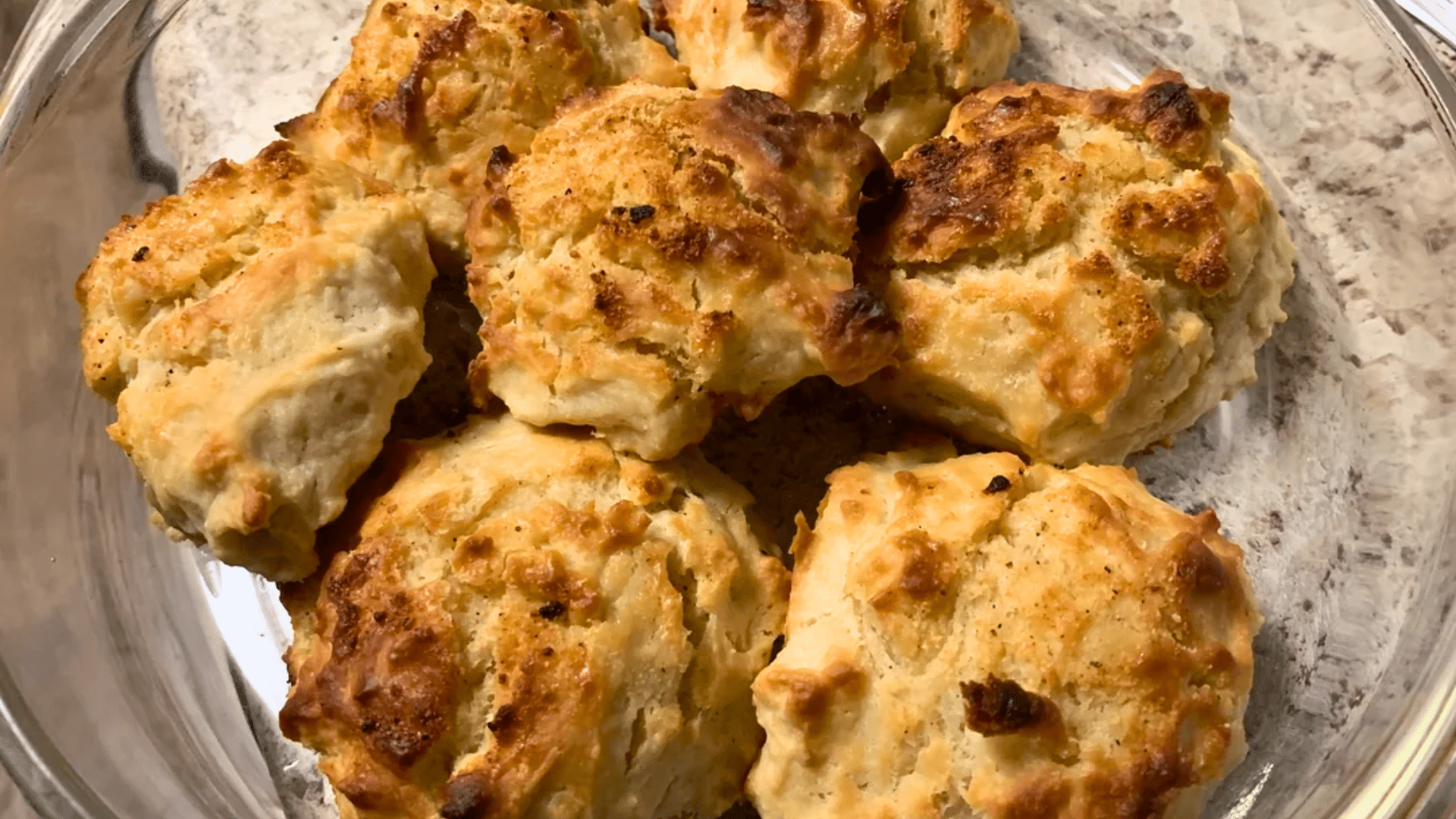 A top-down shot of seven freshly baked, rustic homemade drop biscuits resting in a clear glass baking dish; the tops are unevenly browned and textured, showcasing a hand-crafted, buttery crust.