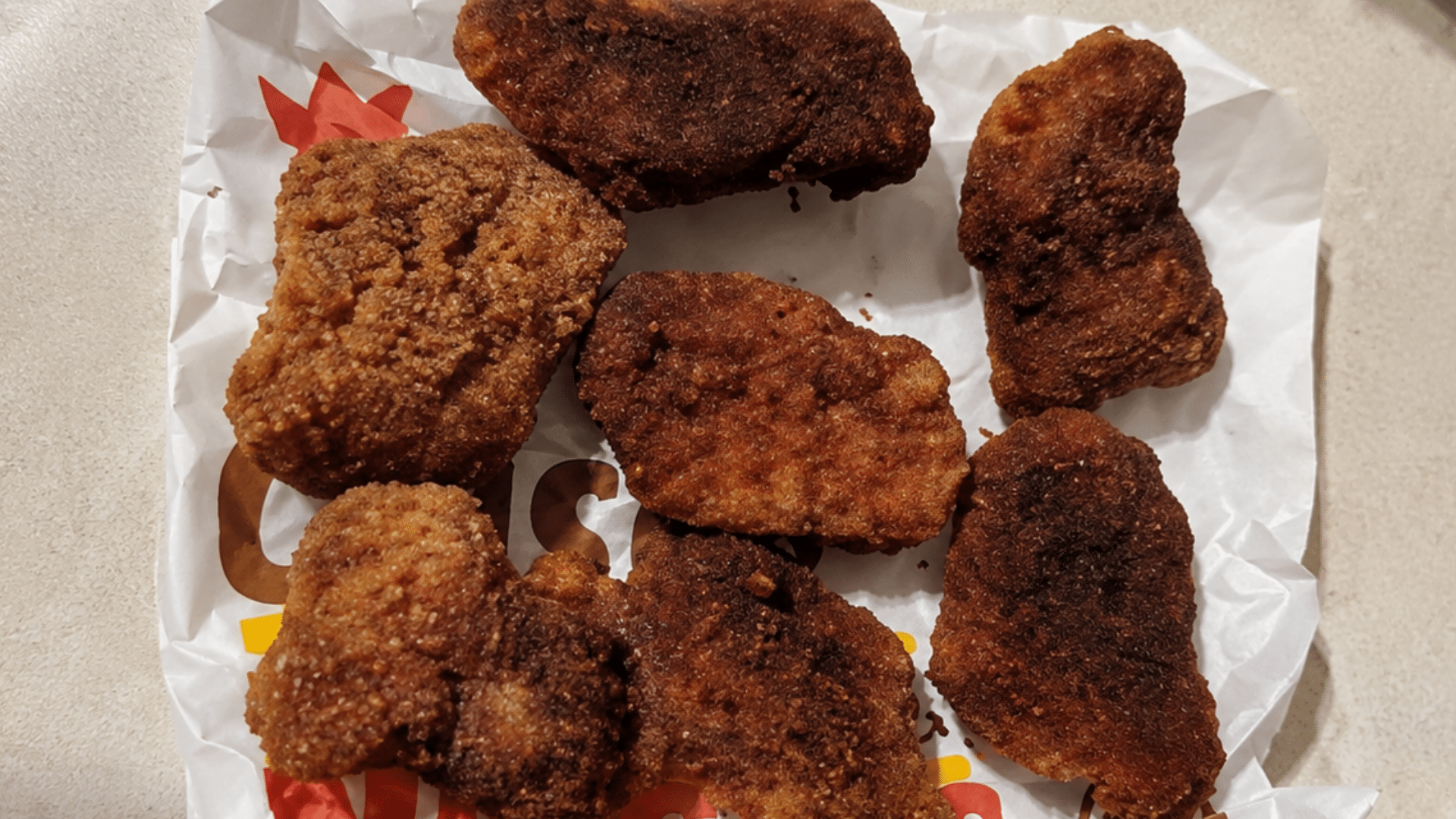 A high-angle shot of seven over-cooked chicken nuggets with a dark, heavily browned and slightly burnt crumb coating resting on a sheet of wrinkled white paper, showcasing an uneven, dry home-baked texture.