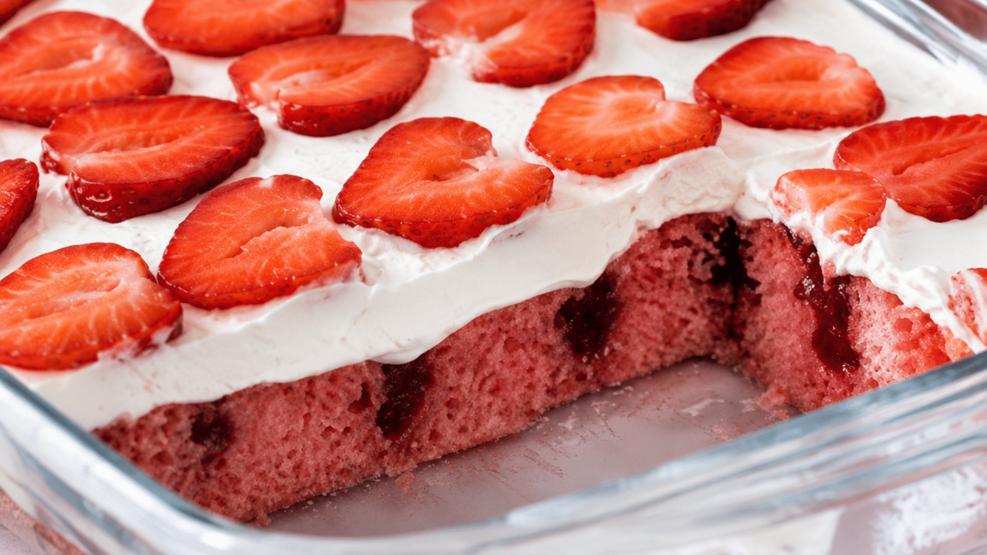 A close-up, high-angle shot of a freshly cut strawberry poke cake in a glass baking dish; the bright pink sponge cake features pockets of darker strawberry glaze, topped with a thick layer of white whipped frosting and neatly arranged heart-shaped strawberry halves.