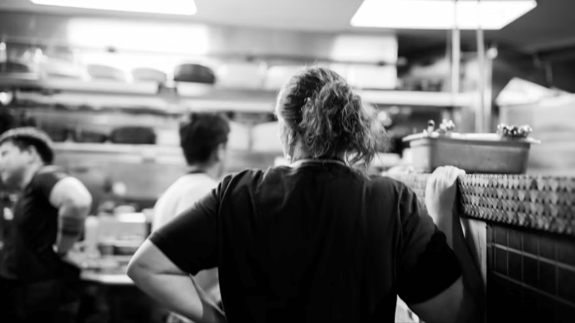 A black-and-white, candid over-the-shoulder shot of a kitchen worker or chef with their hair tied up in a ponytail, standing at a restaurant service counter and looking into a busy, bustling professional kitchen with cookware lining the shelves.