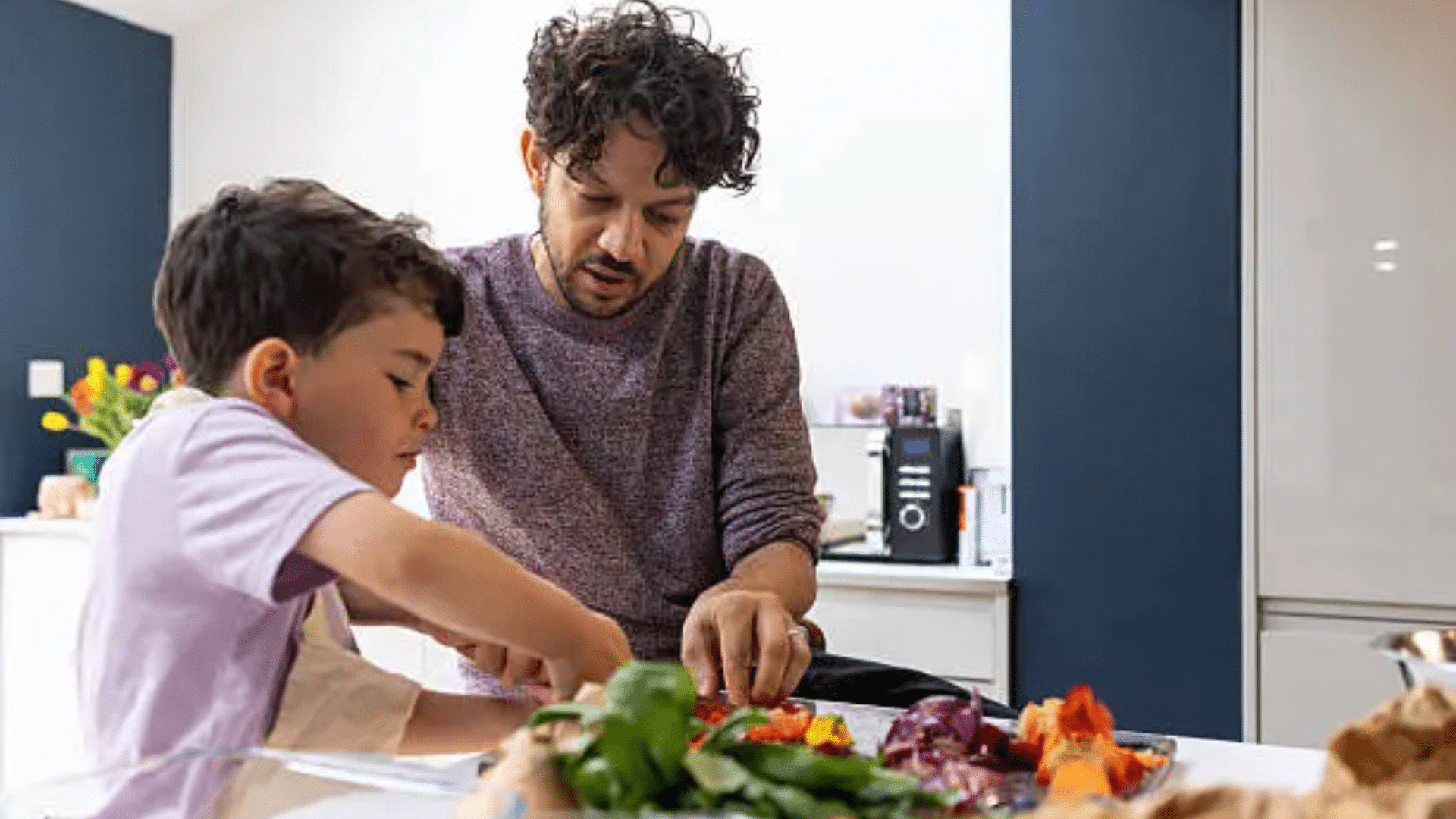A high-angle shot of a father assisting his young son in a kitchen; the boy is focused on chopping vegetables on a metal tray, illustrating a moment of shared learning and positive father-son bonding.