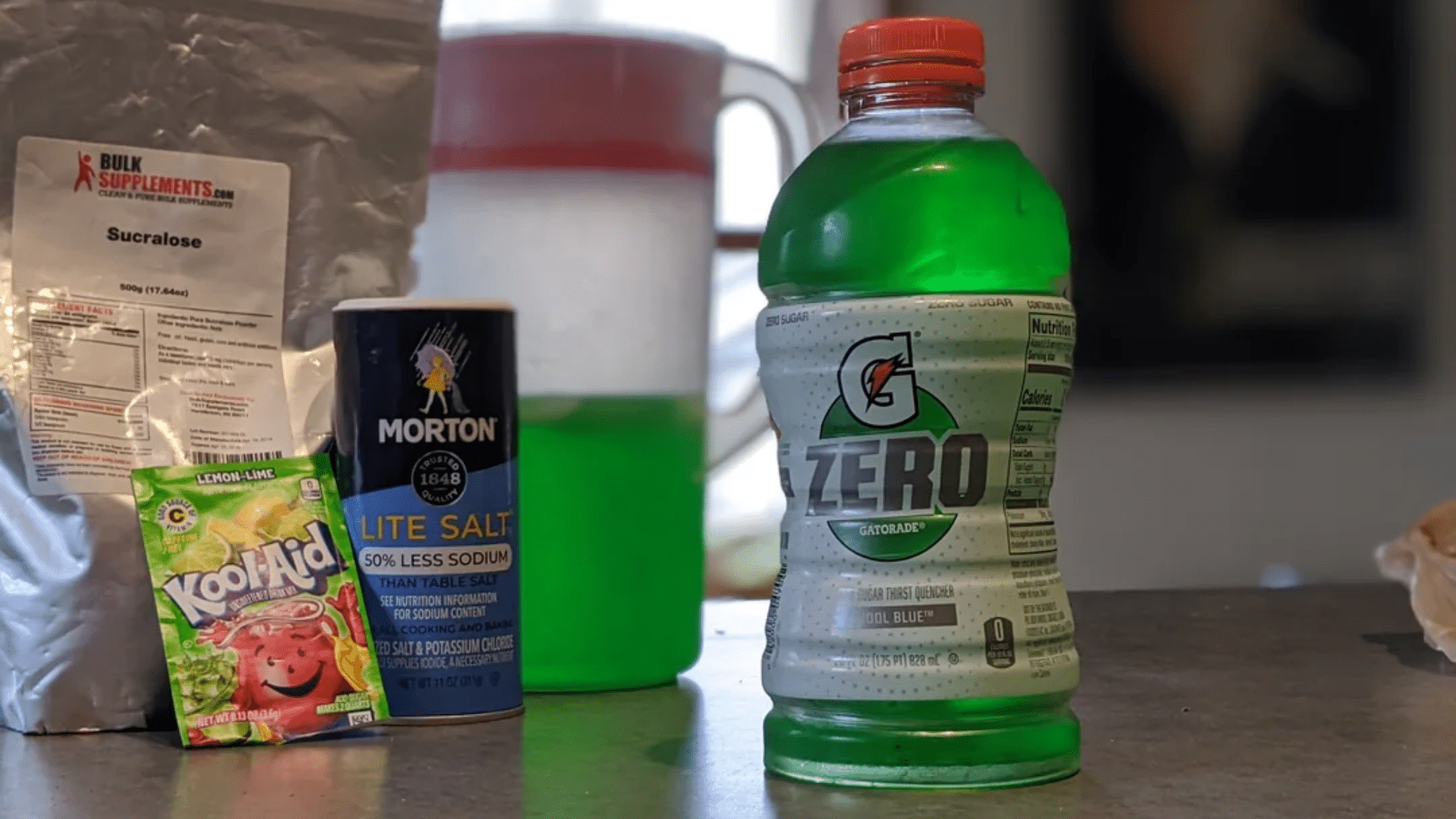 A kitchen counter staging area showing a green Gatorade Zero bottle next to a "Lite Salt" container, a bulk bag of Sucralose, and a Lemon-Lime Kool-Aid packet, representing the DIY "Keto-Aid" trend.