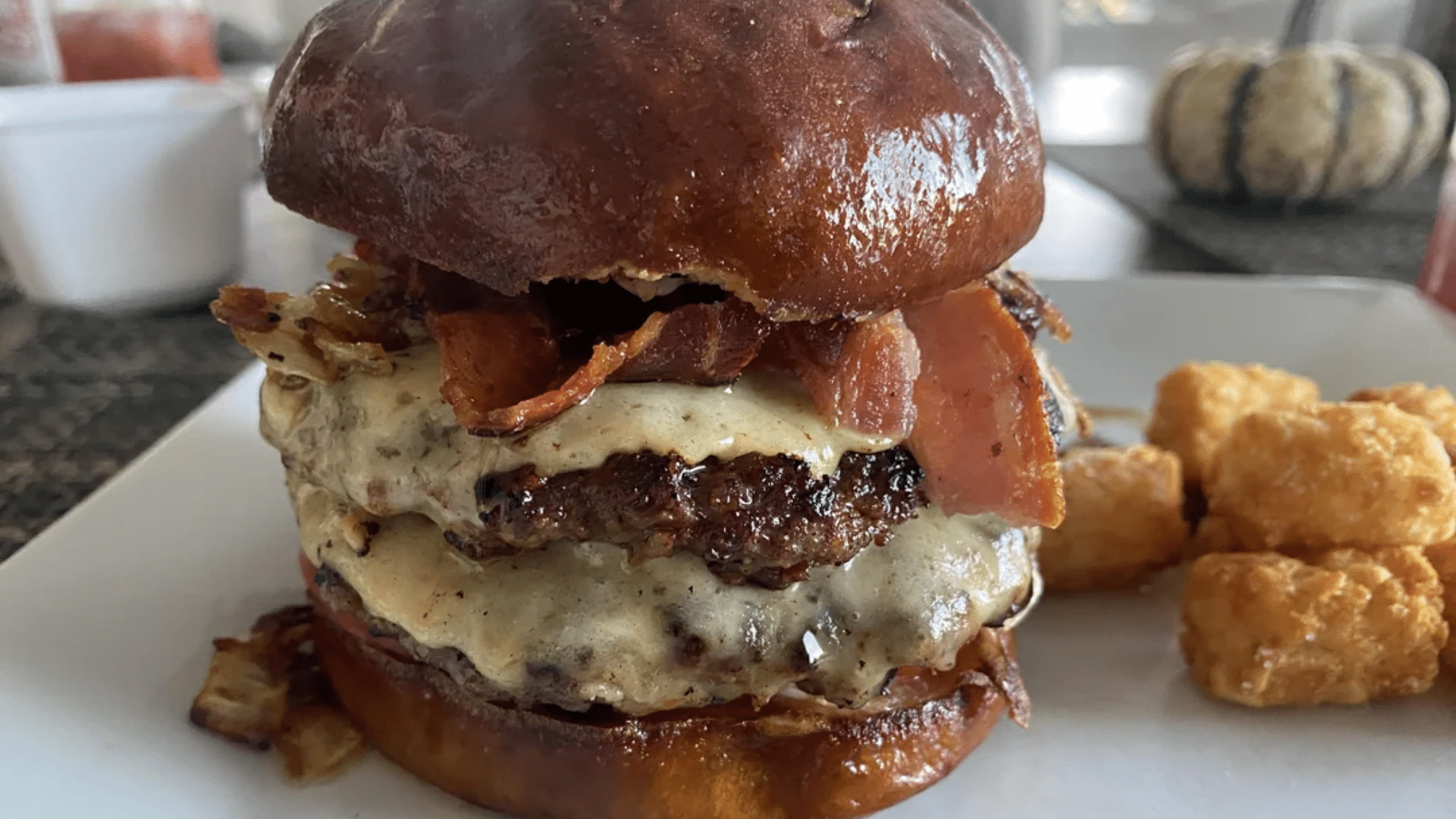 A close-up, tempting shot of a large BBQ Bounty Whopper; the burger features two flame-grilled beef patties, melted Swiss cheese, and thick-cut bacon on a shiny toasted brioche-style bun, served with a side of golden-brown tater tots.
