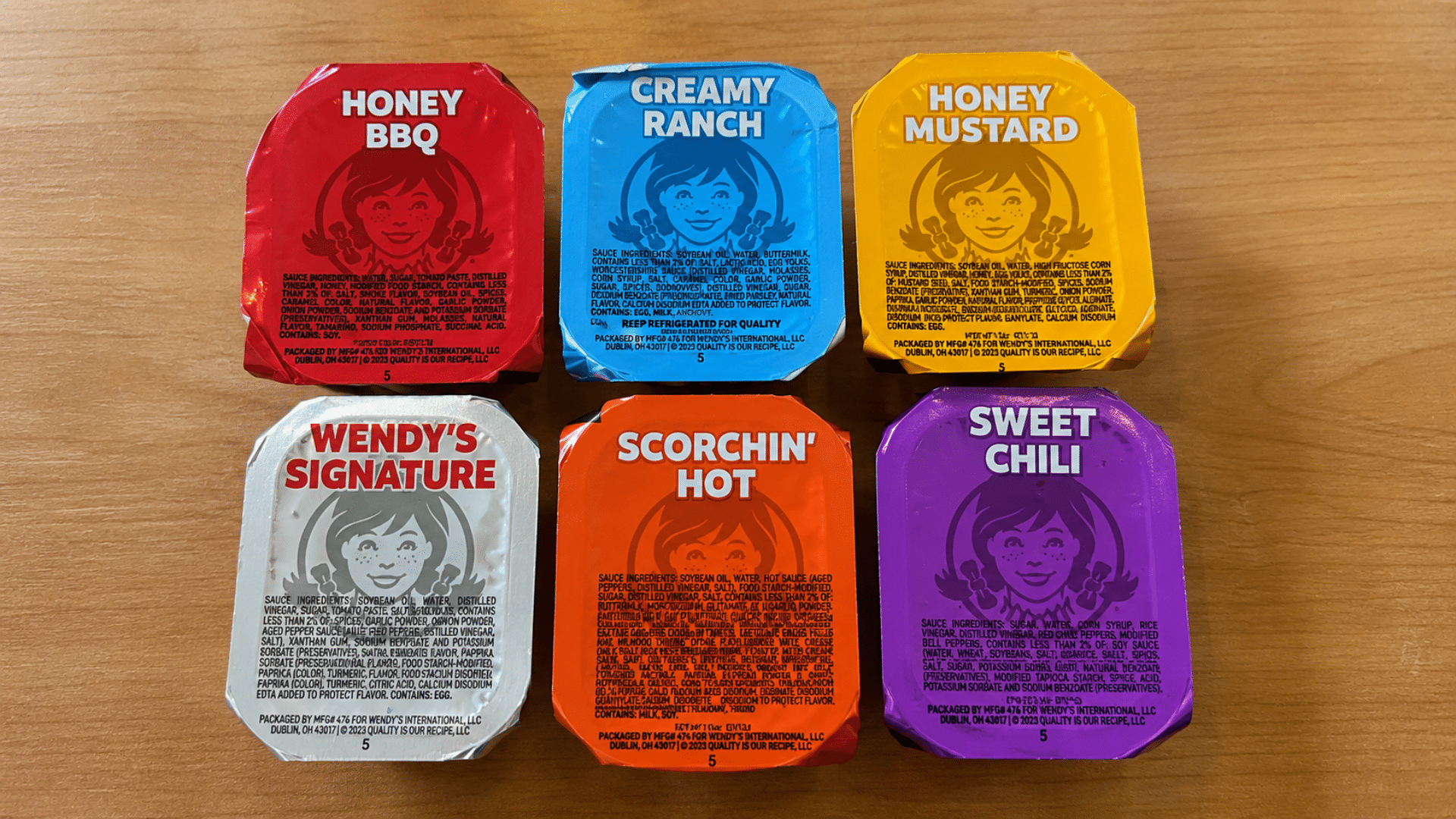 A top-down shot of six rectangular Wendy's dipping sauce containers arranged in two rows; flavors include Honey BBQ, Creamy Ranch, Honey Mustard, Wendy's Signature, Scorchin' Hot, and Sweet Chili, each with its signature color-coded foil lid.