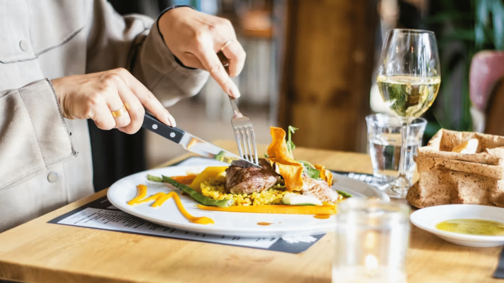 A close-up, high-angle shot of a person using a knife and fork to cut into a plated meal at a restaurant.