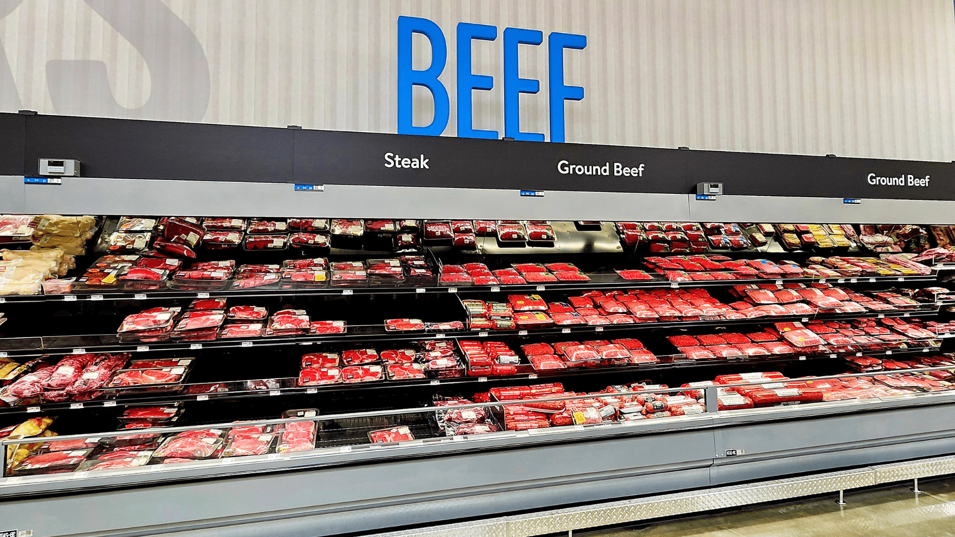 A large blue "BEEF" sign hangs above grocery store refrigerator shelves fully stocked with packaged red meat.