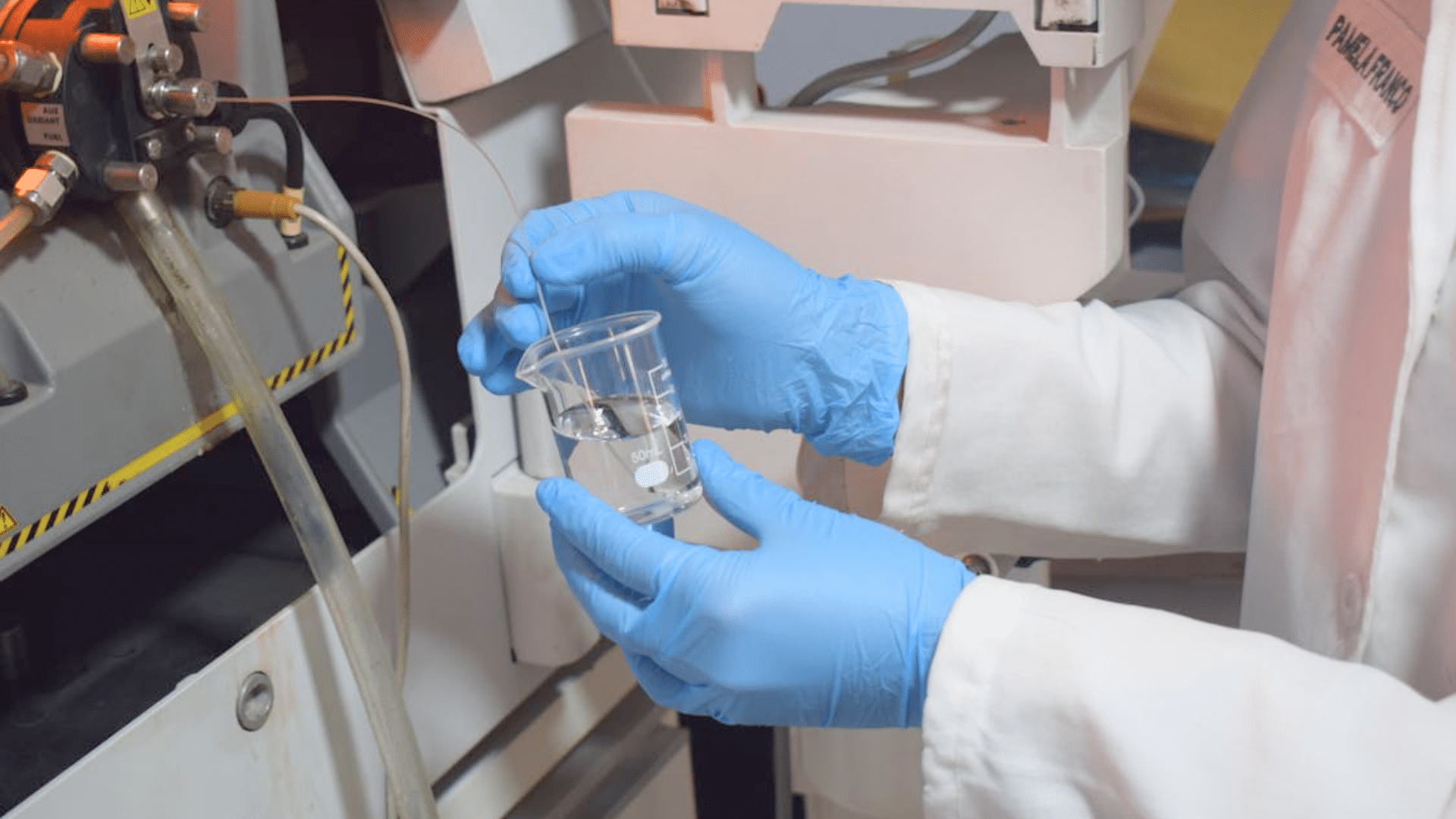 A scientist wearing a lab coat and blue gloves holding a beaker of clear liquid next to laboratory equipment.