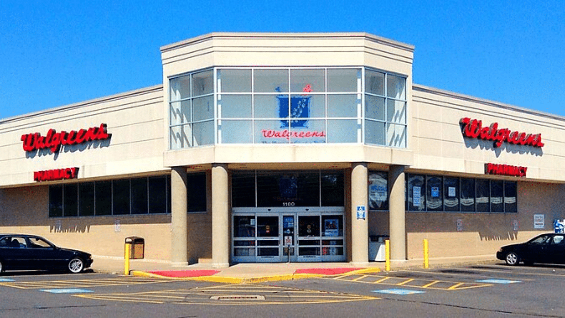 The exterior of a Walgreens pharmacy building with a large glass entryway and red signs on a bright, sunny day.