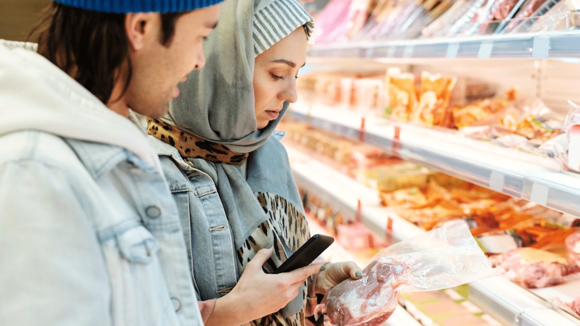 A man and a woman wearing a hijab look at a smartphone and a package of beef in a grocery store aisle.