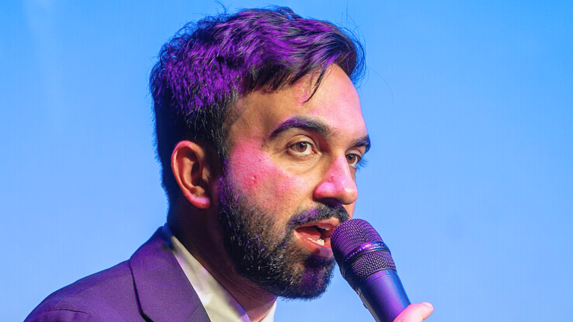 A close-up portrait of New York City mayor Zohran Mamdani speaking into a microphone against a blue background.
