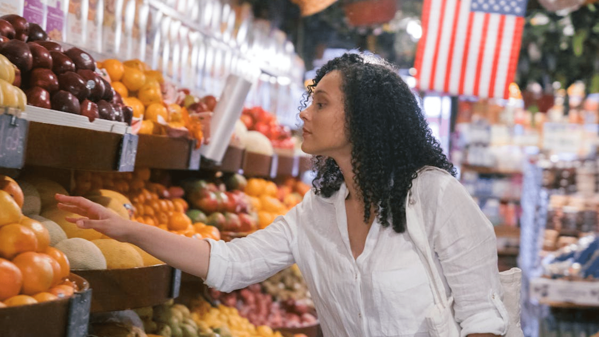 A woman in a white shirt reaches for fresh fruit in a grocery store produce section under a hanging American flag.