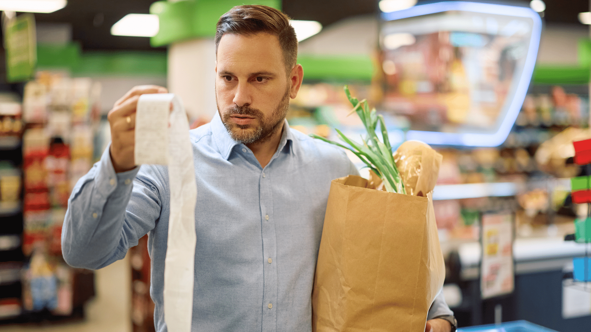 A man holding a brown paper bag full of groceries looks closely at a long receipt inside a supermarket.
