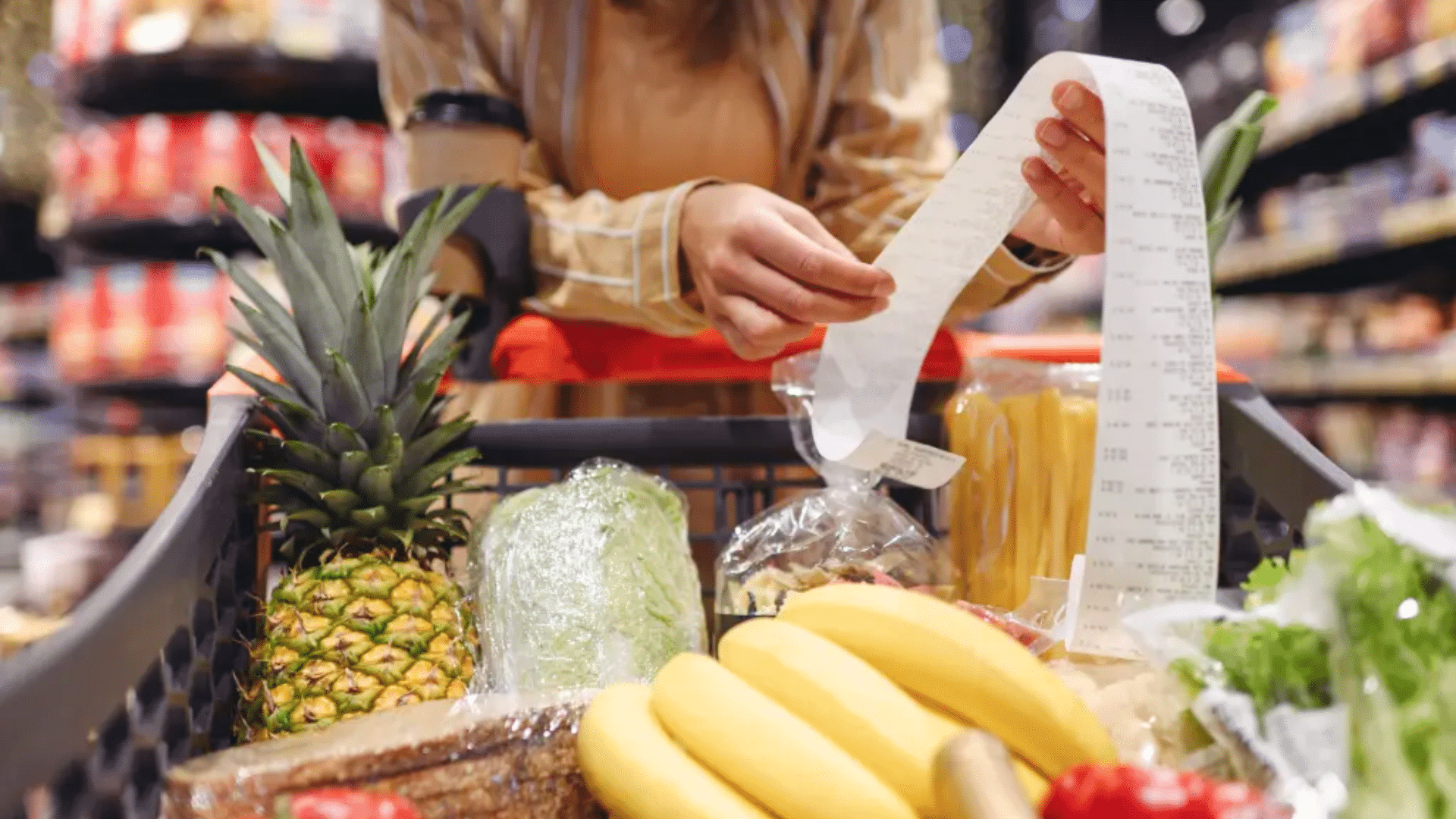 A person looking over a long receipt next to a shopping cart filled with fresh produce like a pineapple and bananas.