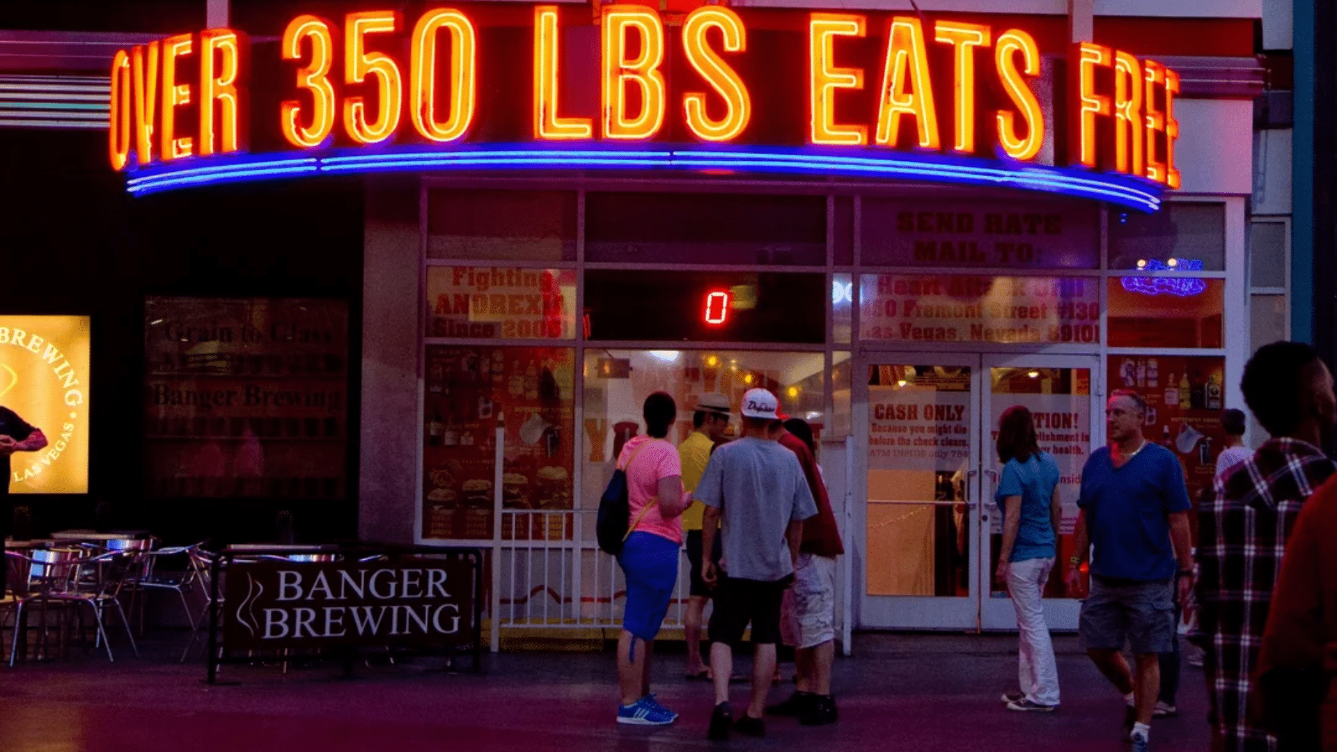 Visitors standing outside Heart Attack Grill beneath a neon “over 350 lbs eats free” sign.