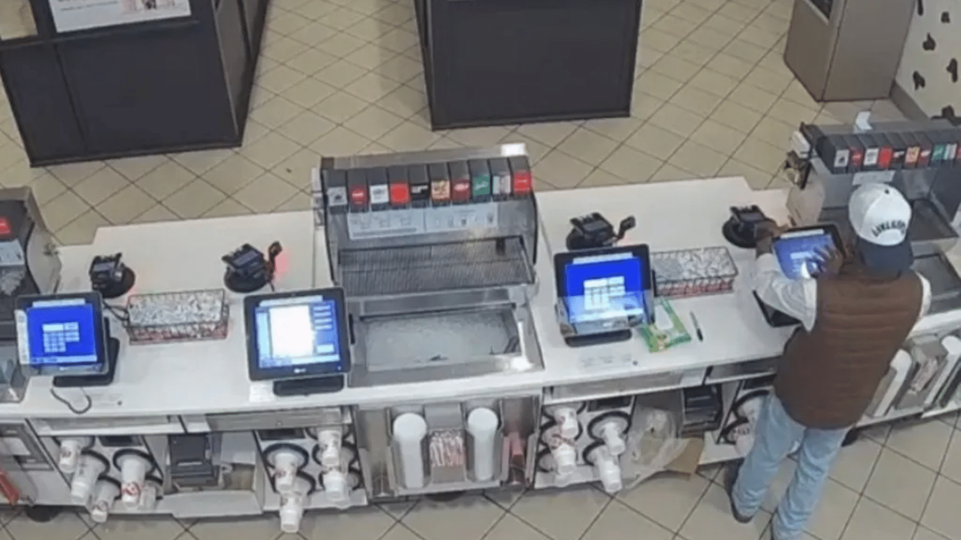 An overhead security camera view shows a person standing at a cash register in an empty fast-food restaurant.
