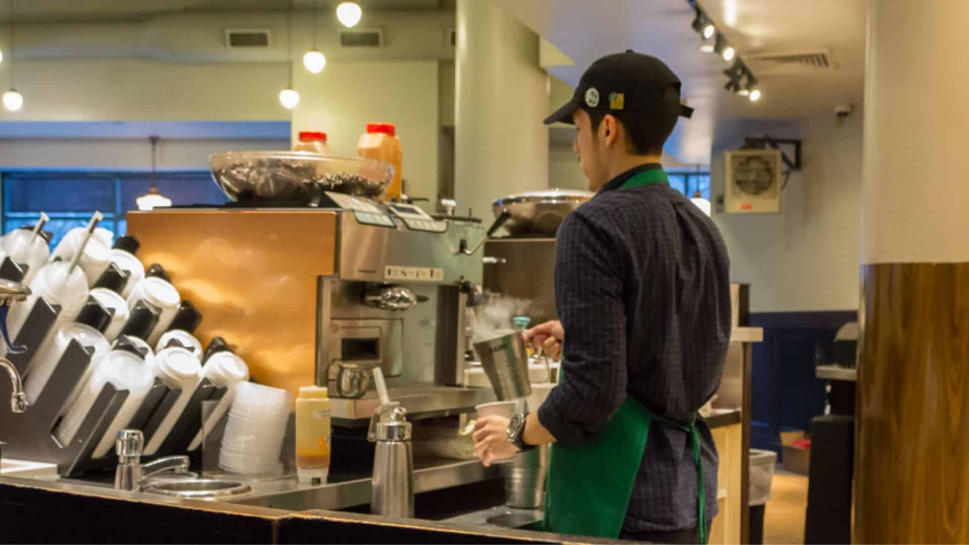 Starbucks employee wearing a green apron prepares drinks behind the counter inside a coffee shop.