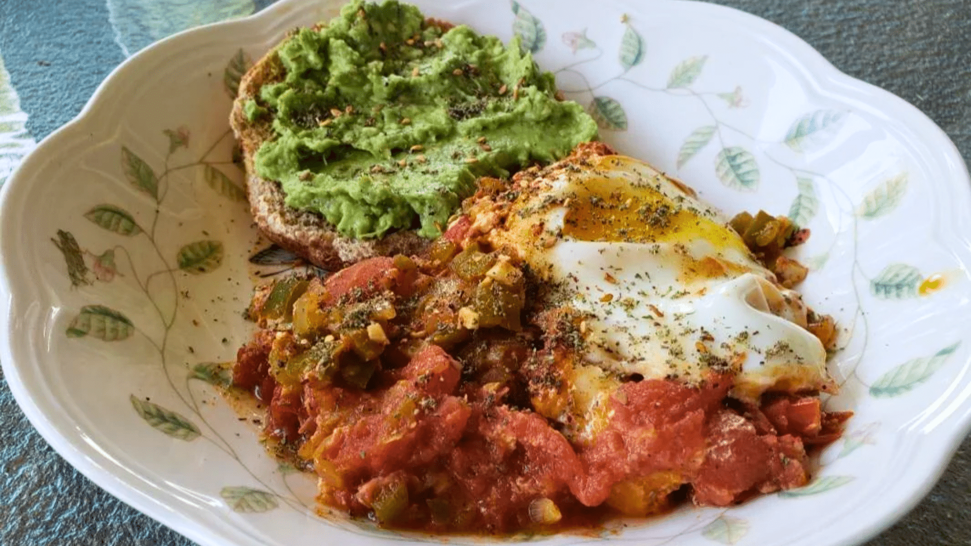 A beautiful plate featuring a slice of toasted artisanal bread spread with vibrant green smashed avocado, served alongside a rich, red Shakshuka tomato-and-pepper stew topped with a perfectly poached egg and sprinkled with dried herbs.