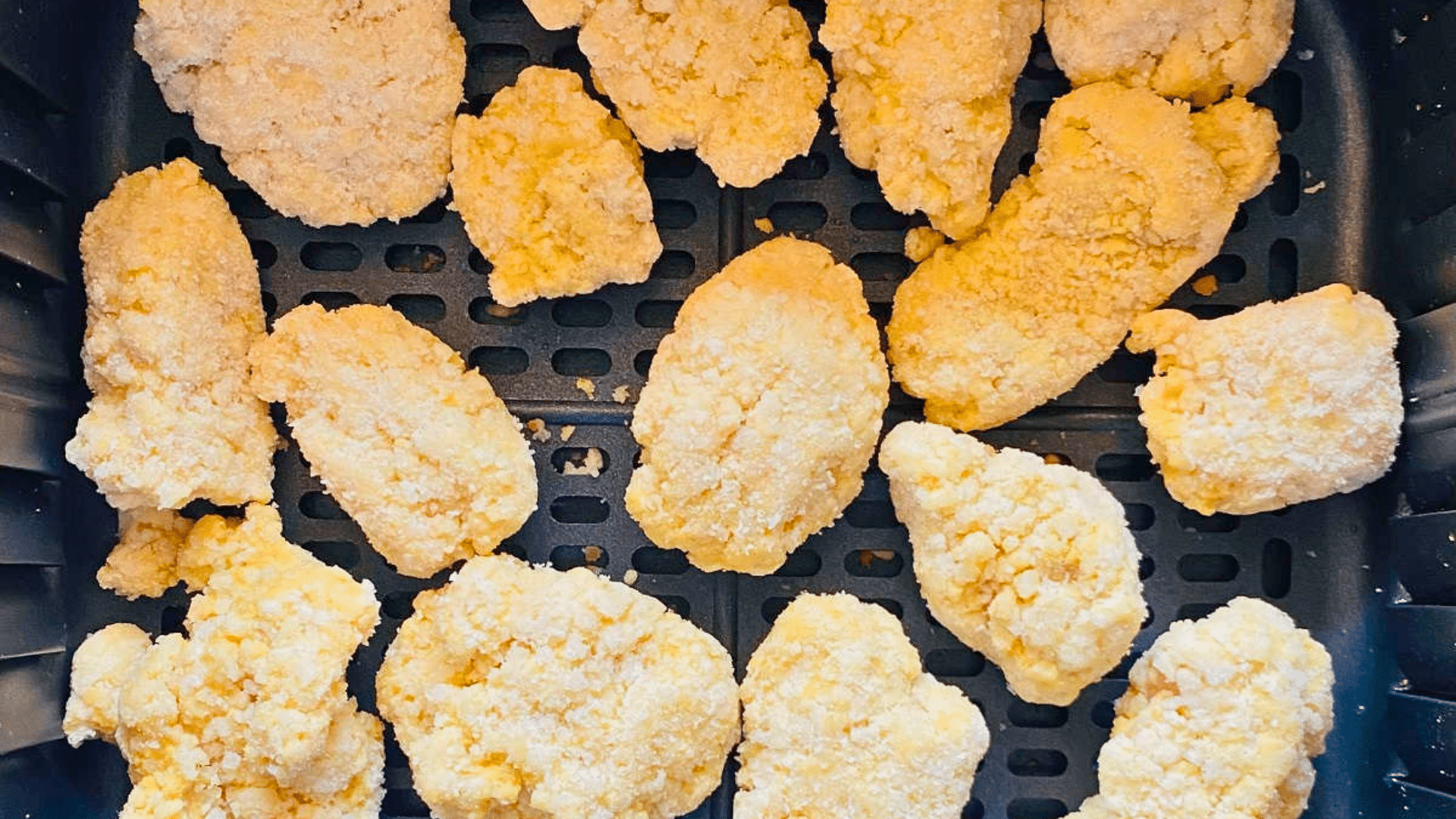 A clean top-down shot looking into a black air fryer basket neatly lined with over a dozen pale, unbaked frozen chicken nuggets coated in a light white flour or cornstarch breading, ready to be cooked.