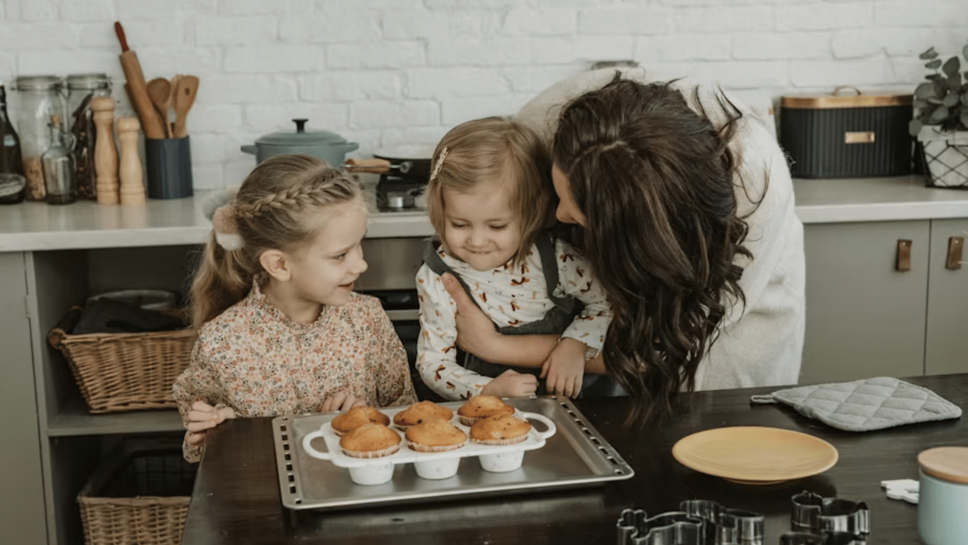 A medium shot of a mother leaning over a kitchen island to gently interact with her two young daughters; a white ceramic baking pan filled with six freshly baked, golden-brown muffins or cupcakes sits on a metal baking tray on the dark wooden table.