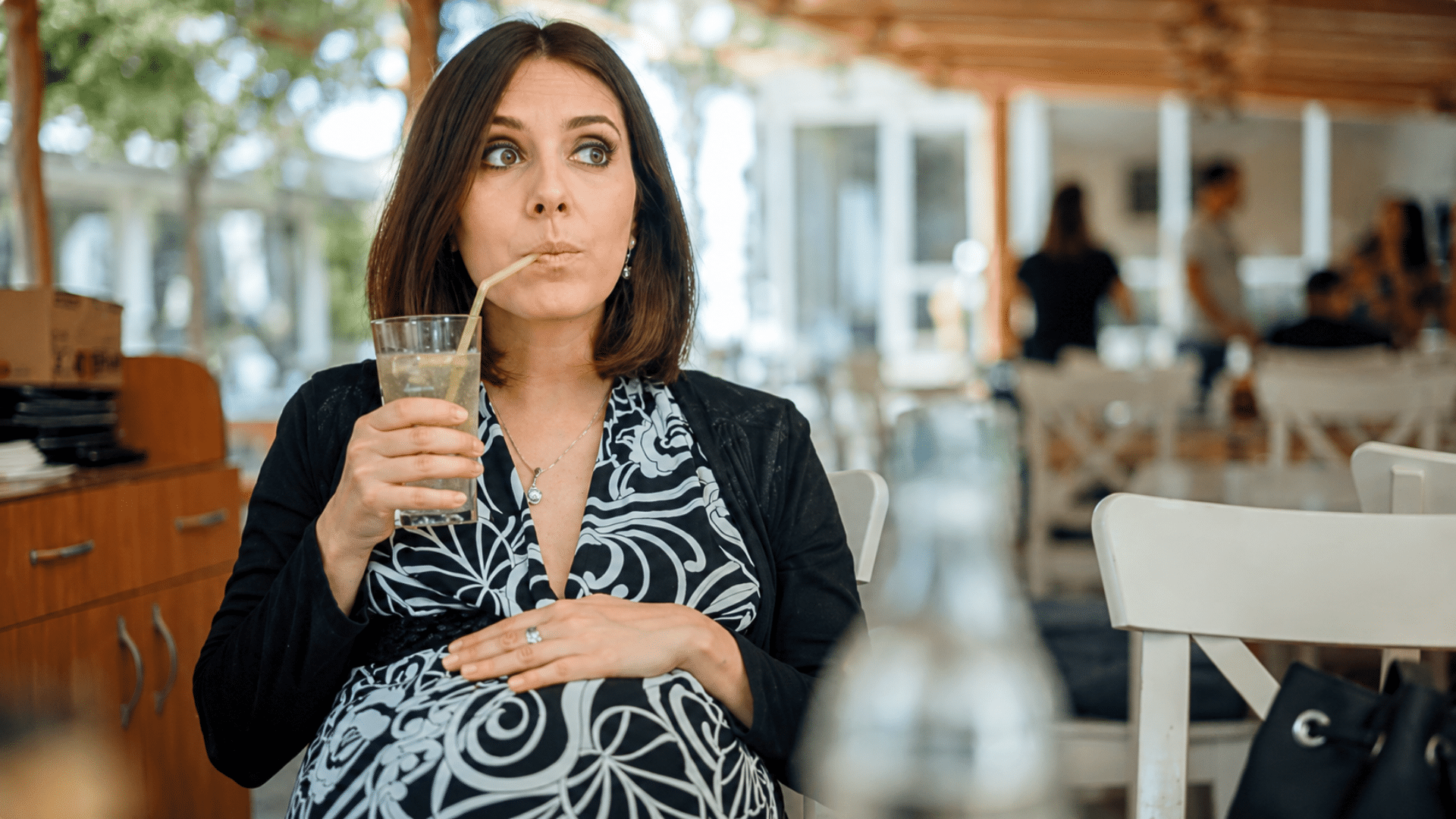 A medium shot of a pregnant woman sitting in a restaurant terrace, cradling her baby bump while drinking a refreshing iced beverage with a straw; she looks off to the side with an expressive, pleasantly surprised look.