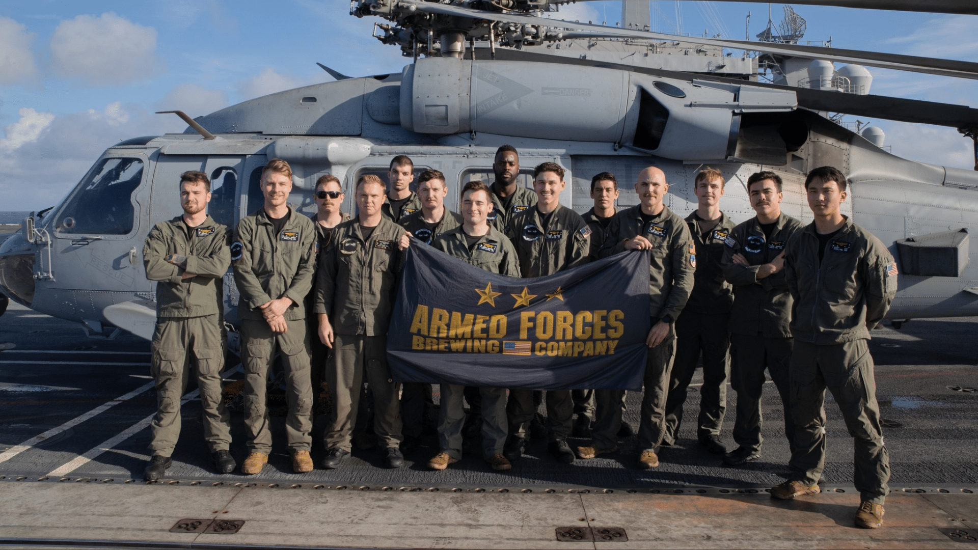 A group of military personnel in flight suits standing on an aircraft carrier flight deck in front of a helicopter, holding up a black "Armed Forces Brewing Company" banner.