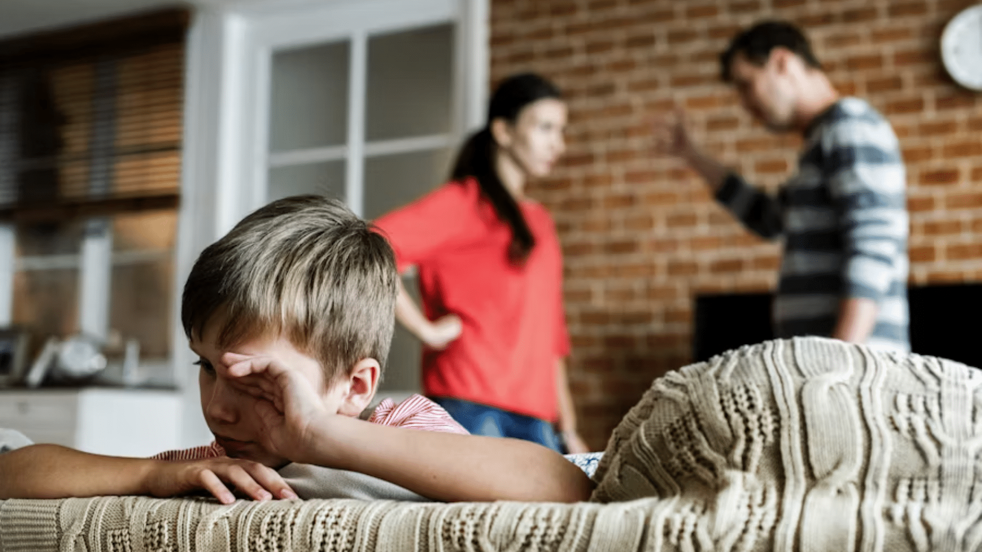 A dramatic shot from behind a young boy sitting on a sofa, rubbing his eye as if crying; in the blurred background, his parents are engaged in a heated standing argument in a brick-walled living room.