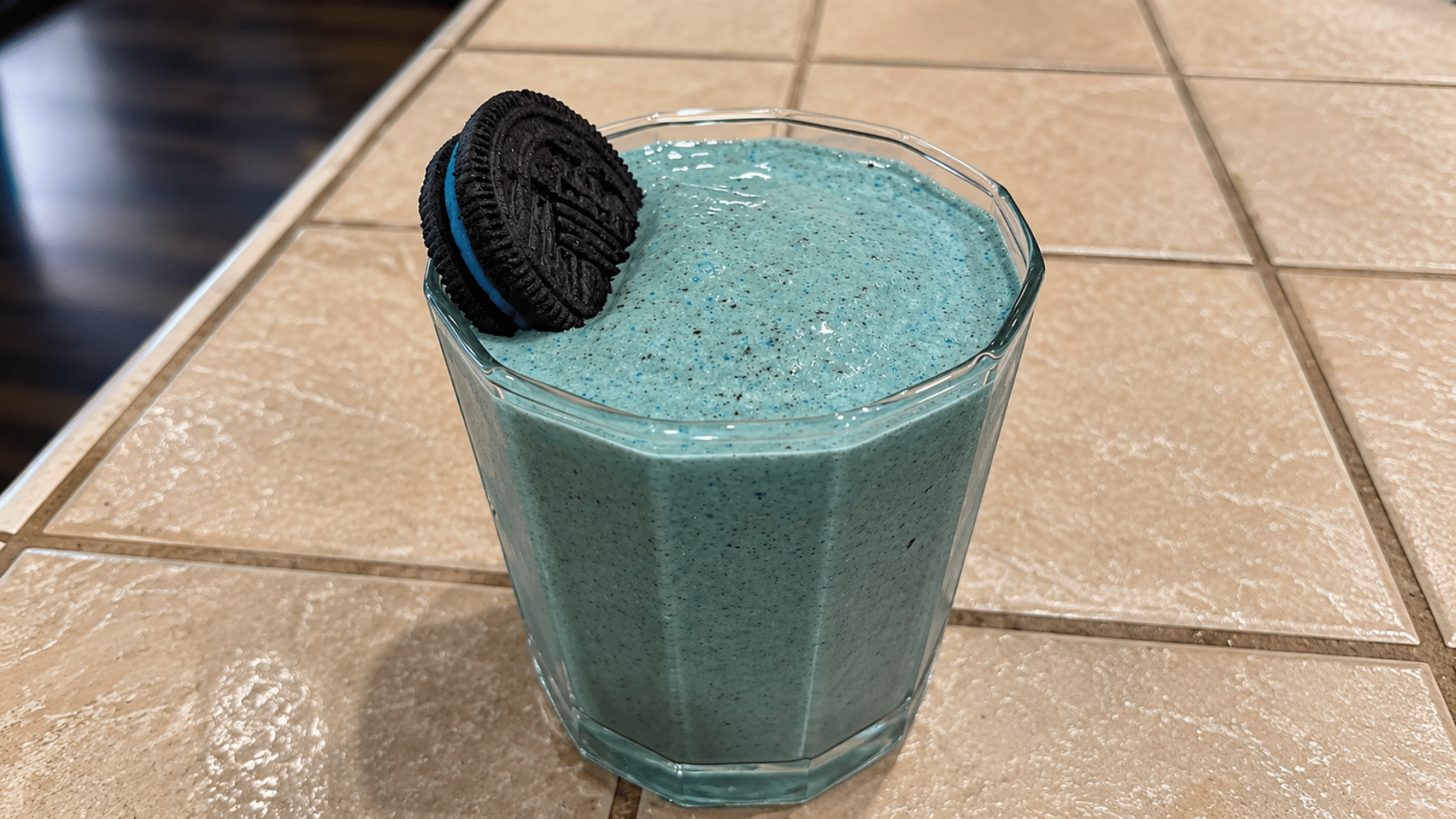 A close-up of Grogu's Blue Cookie Shake sitting on a tan tiled counter; the creamy, bright blue milkshake is topped with a whole dark chocolate sandwich cookie.
