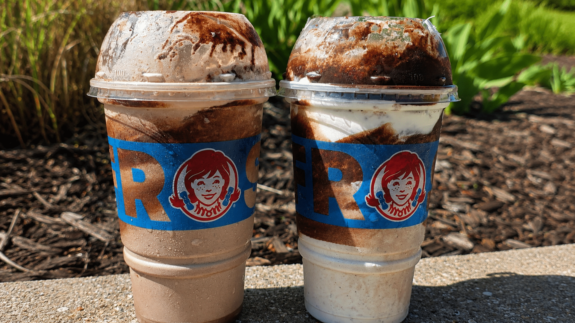 A close-up of two large Frosty Swirls™ sitting on a concrete ledge outdoors; the left is a chocolate-based swirl with dark chocolate sauce streaks, and the right is a vanilla-based swirl with the same dark chocolate drizzle, both under domed lids.