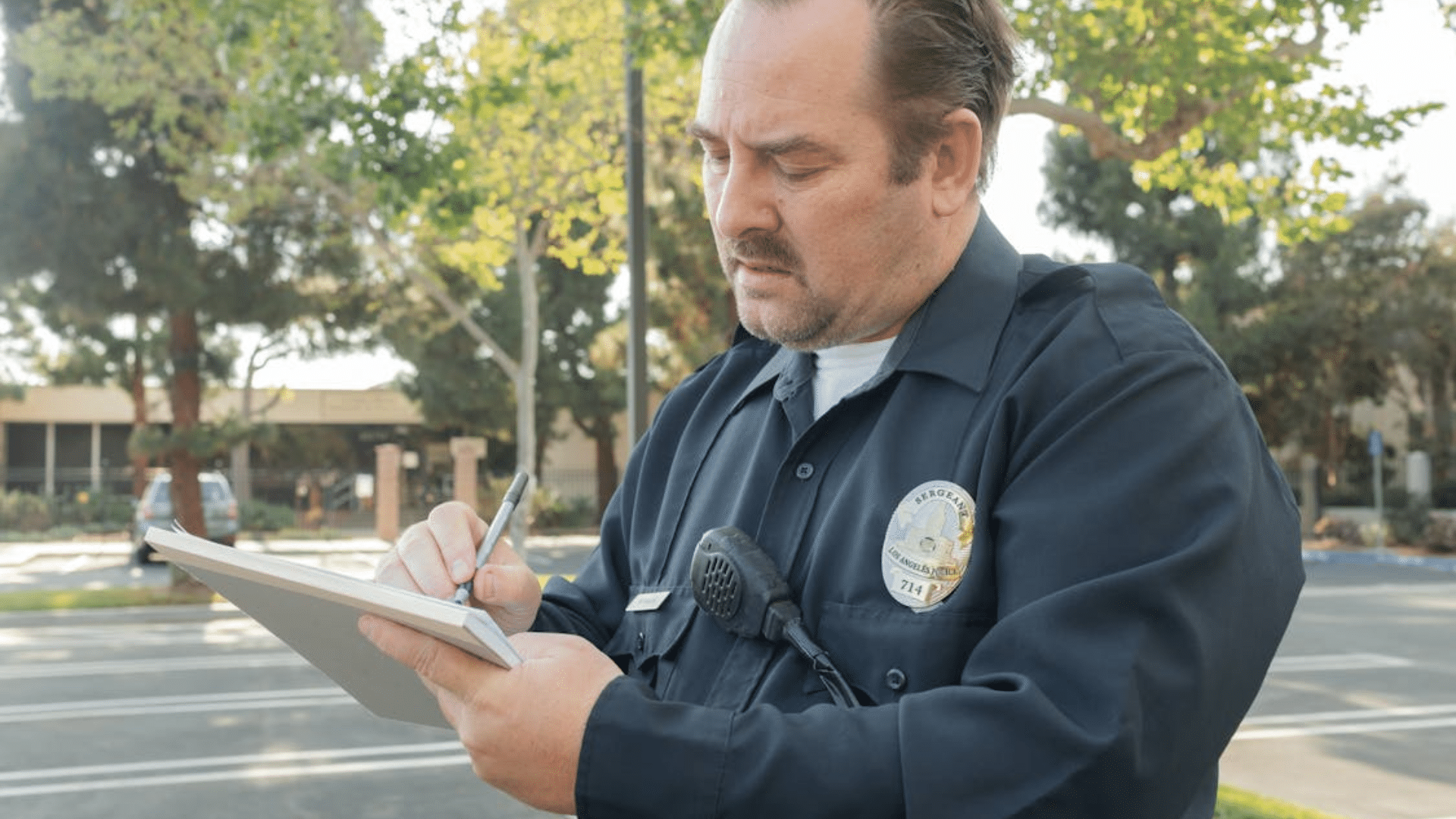 A male police officer in a dark blue uniform writes on a notepad while standing outside on a sunny day.