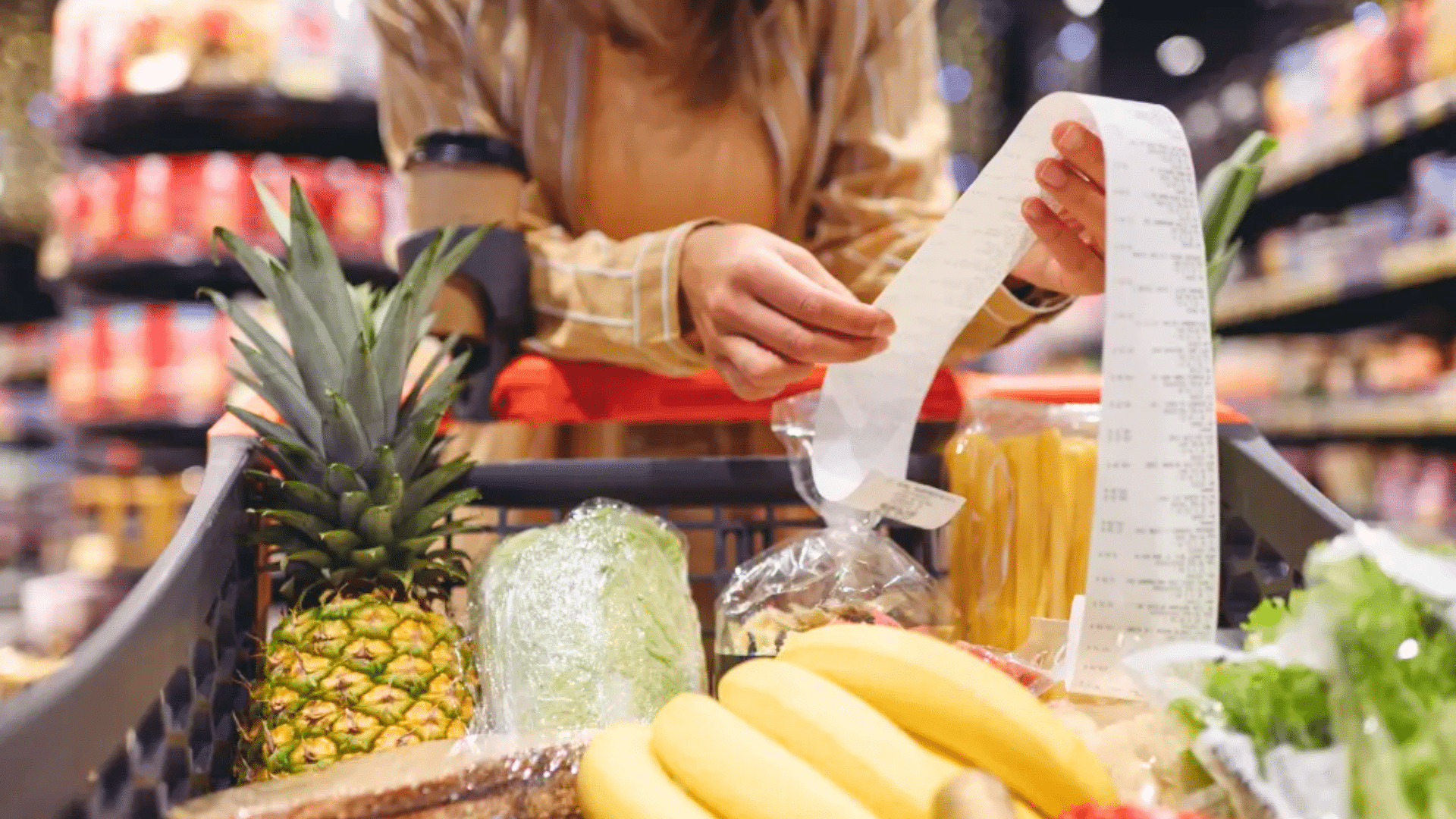 A person looking over a long receipt next to a shopping cart filled with fresh produce like a pineapple and bananas.