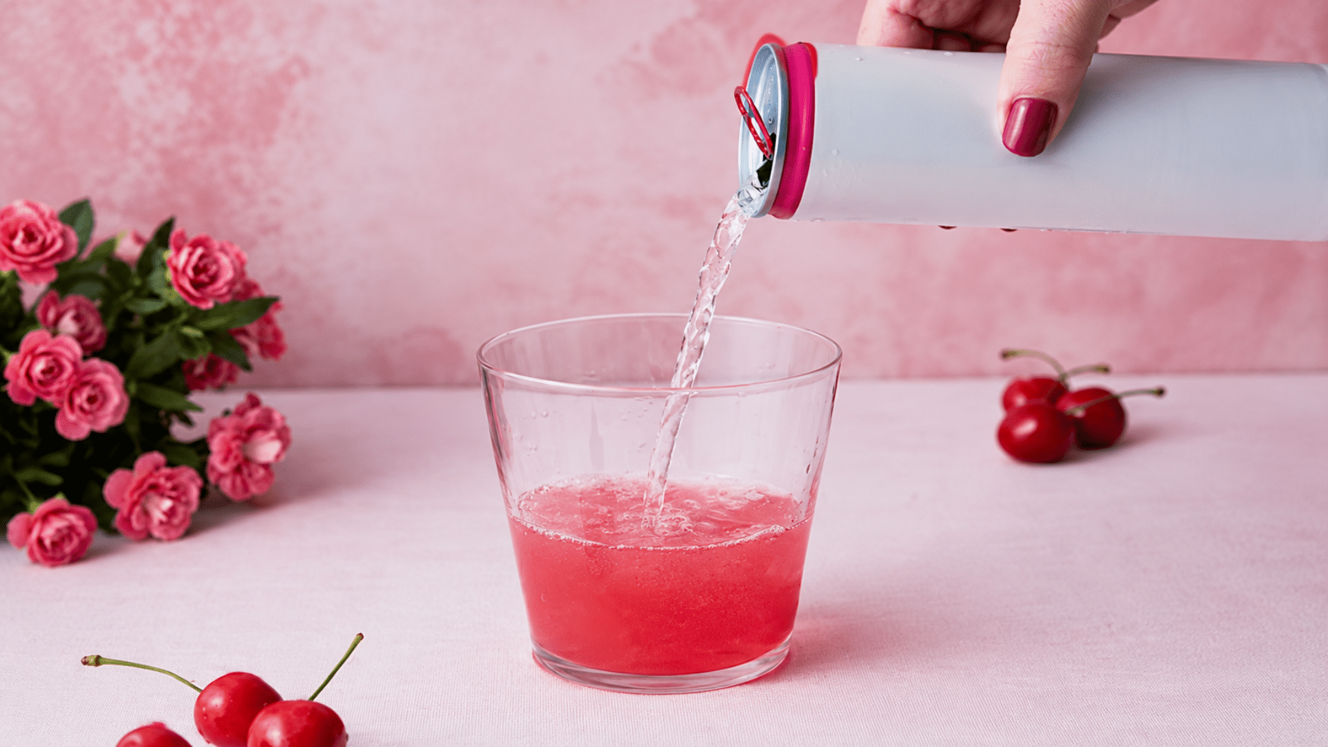 Pink beverage being poured into a glass with cherries on a table nearby.