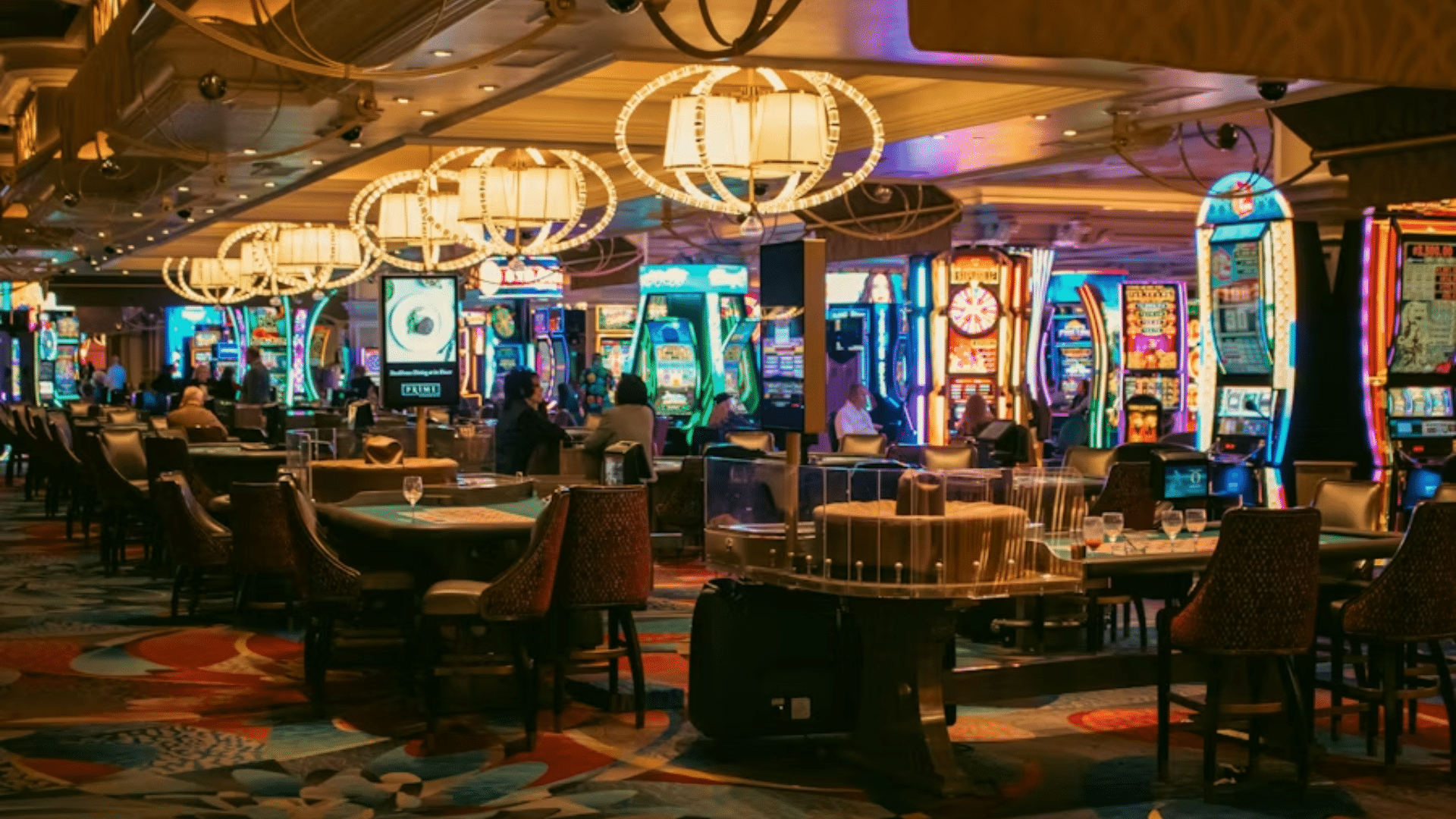 Casino floor with illuminated slot machines and tables under chandelier lighting.