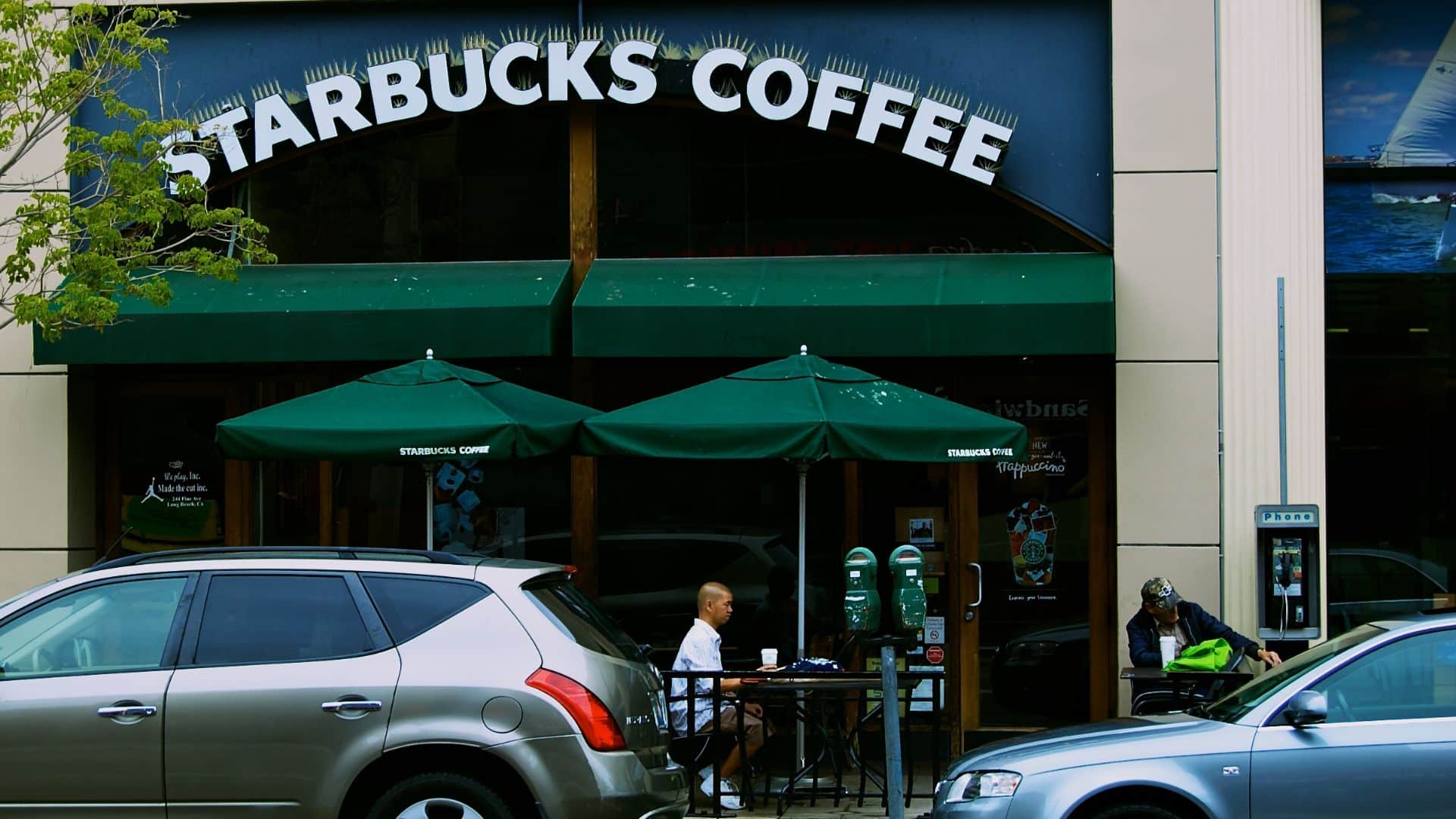 Exterior of a Starbucks Coffee storefront with green umbrellas, parked cars, and customers seated outside.