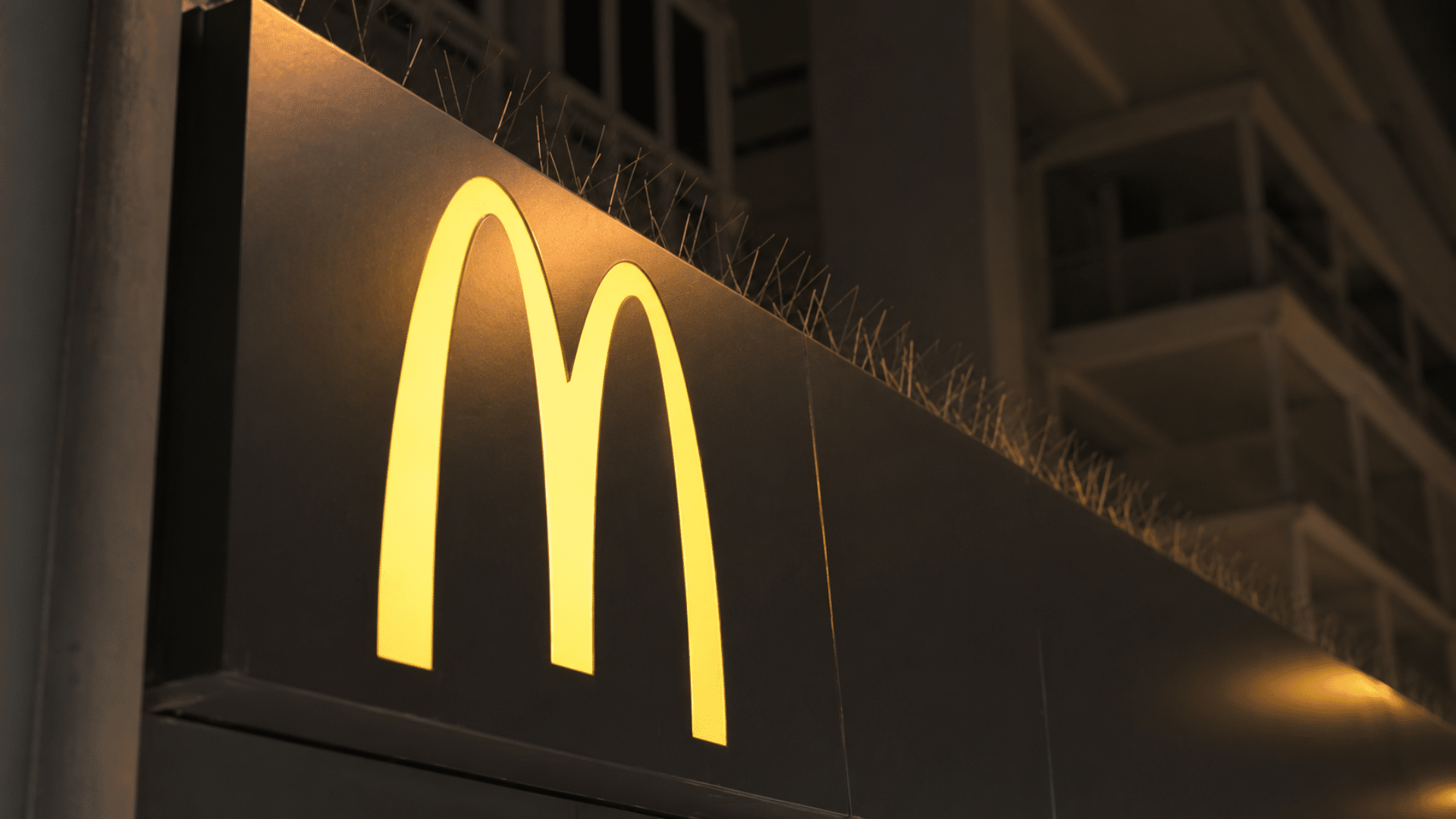 A low-angle shot taken at night of a sleek, dark corporate facade featuring a glowing yellow McDonald's golden arches logo mounted on a black architectural panel, with anti-bird spikes visible along the top edge.