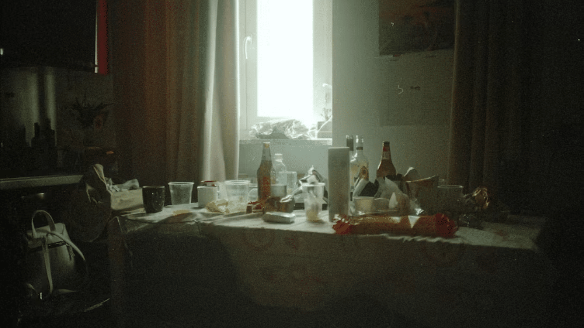 A low-light, moody interior shot of a messy dining table covered in plastic cups, soda and beer bottles, paper plates, and plastic food wrappers after a party or dinner gathering, backlit by a bright, hazy window in the background.