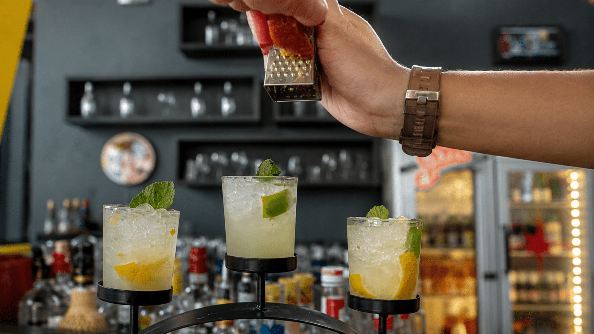 A close-up shot of three citrus-infused ginger ale mocktails garnished with mint leaves, arranged neatly on a curved black metal serving stand at a bar; a bartender's hand is visible in the upper frame using a small metal grater to zest a garnish over the drinks.