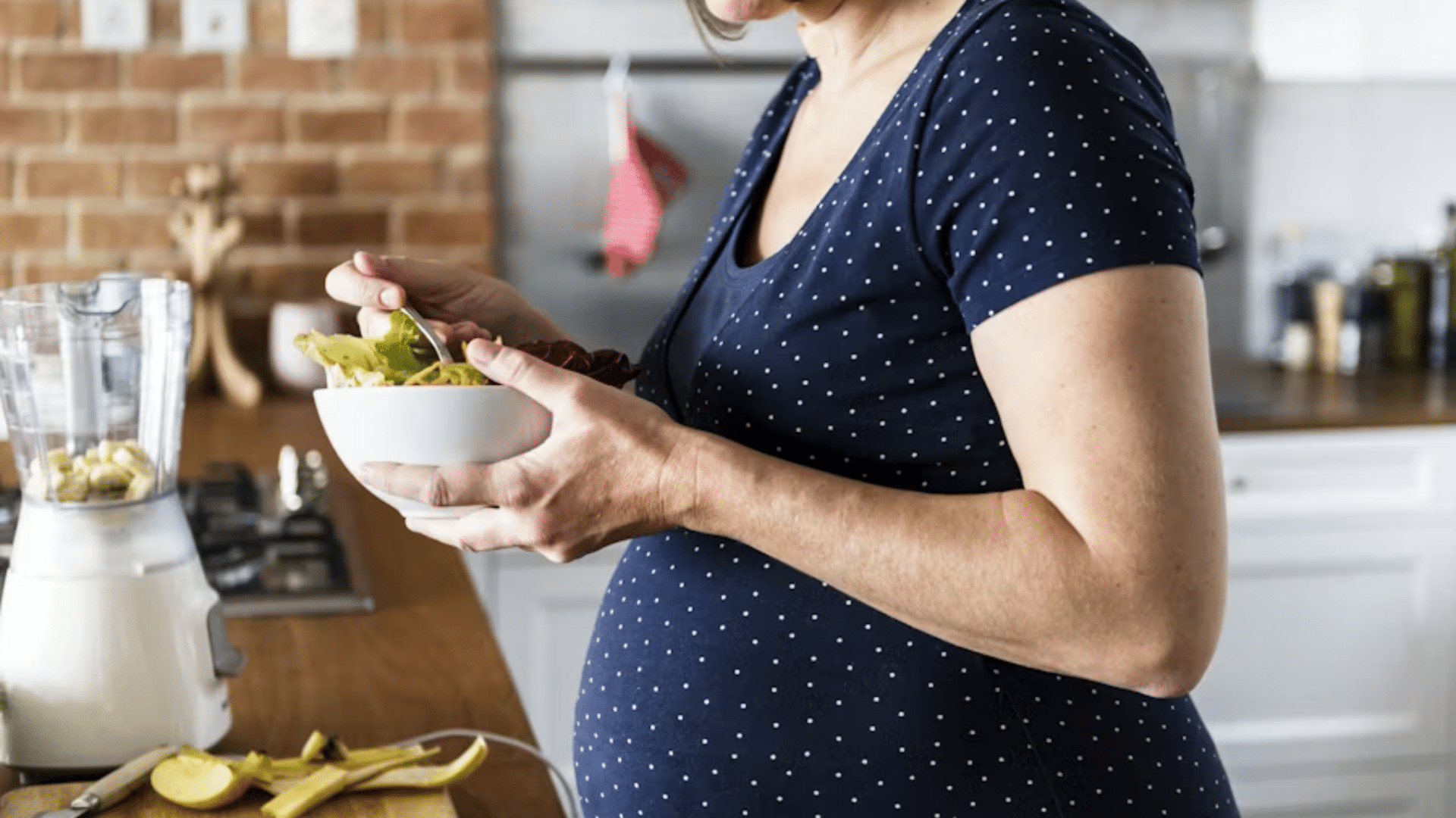 A profile shot of a pregnant woman in a navy blue polka-dot t-shirt standing in a kitchen, holding a white bowl of fresh mixed salad leaves and carrots while preparing a healthy meal next to a blender.