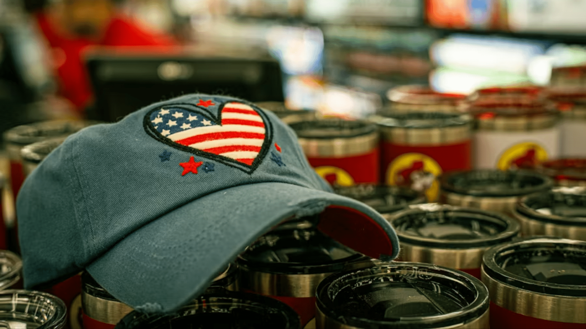 A blue baseball cap featuring an embroidered heart-shaped American flag patch, resting on a retail shelf display of stainless steel travel tumblers.