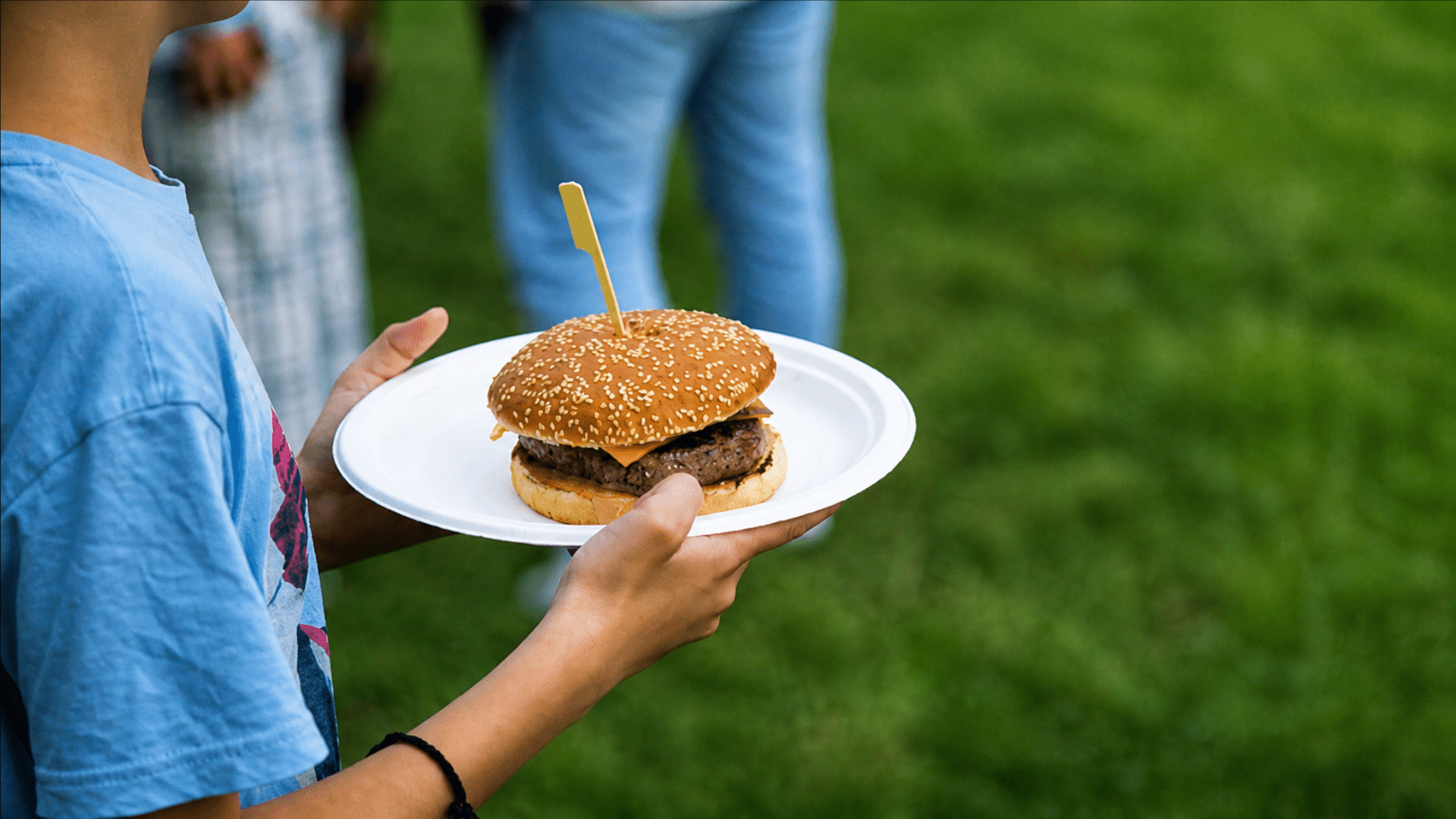 A close-up of a child's hands holding a white paper plate with a large cheeseburger on a sesame seed bun; the background shows a blurred green lawn and other guests.