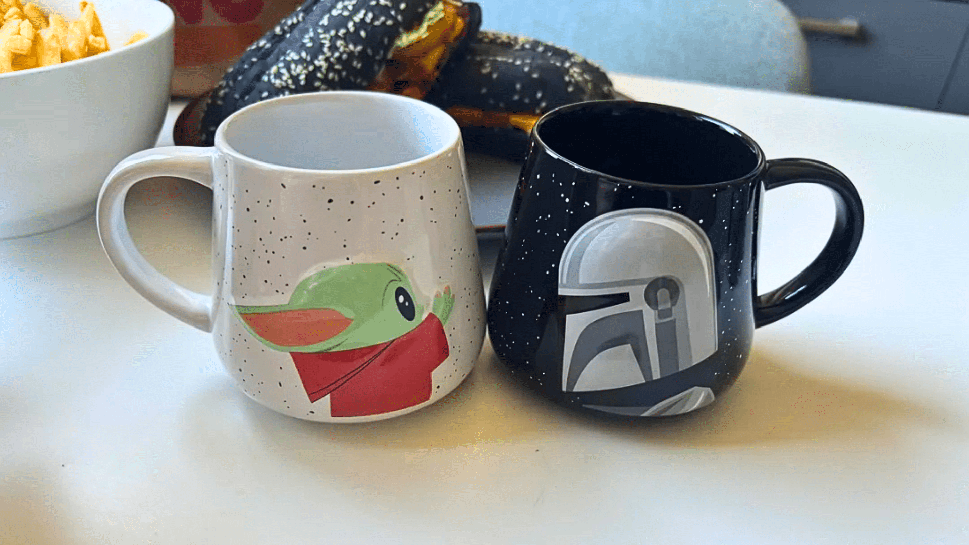 A close-up of two collectible ceramic or plastic mugs sitting on a white table; one is white with black specks featuring an illustration of Grogu in his red robe, and the other is black with white "stars" and a detailed grey Mandalorian helmet graphic.