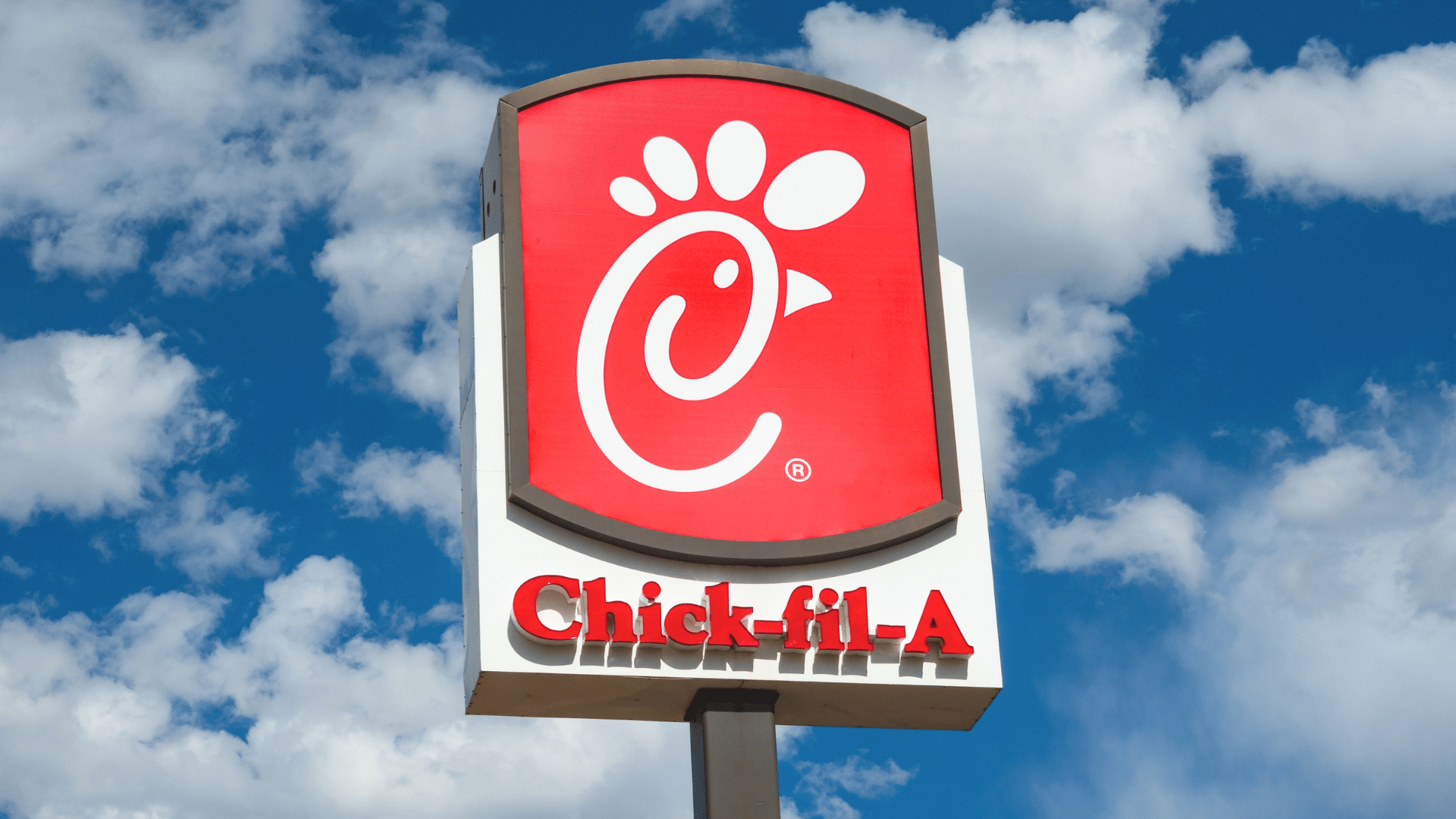 A large red and white Chick-fil-A sign stands tall on a pole against a bright blue sky with white clouds.