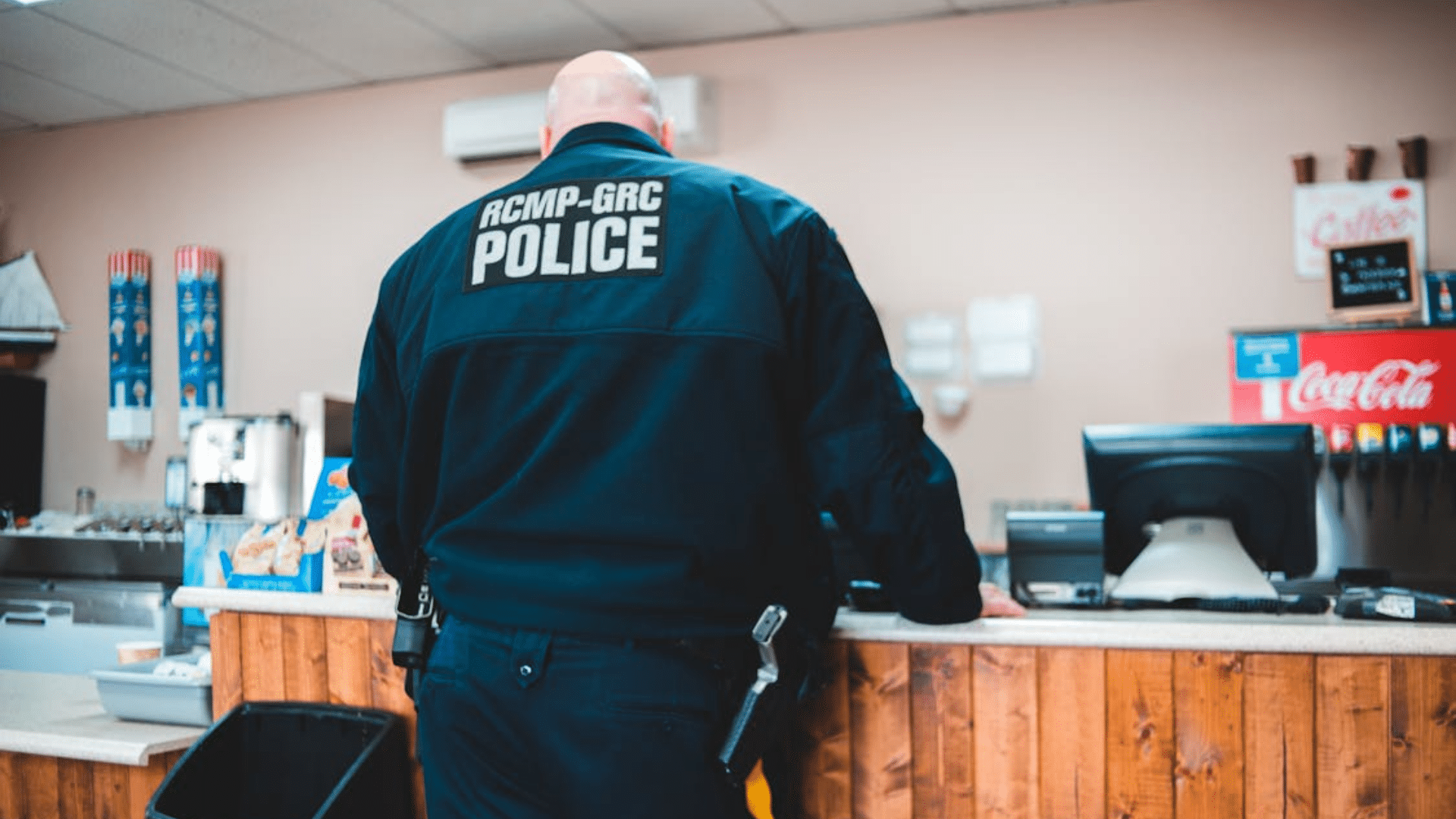 The back of a bald police officer wearing a dark RCMP-GRC POLICE jacket standing at a counter inside a diner.