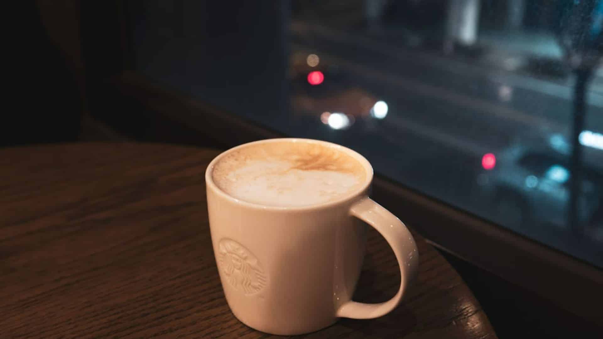 A warm cappuccino on a wooden table by a window overlooking a city street at night.