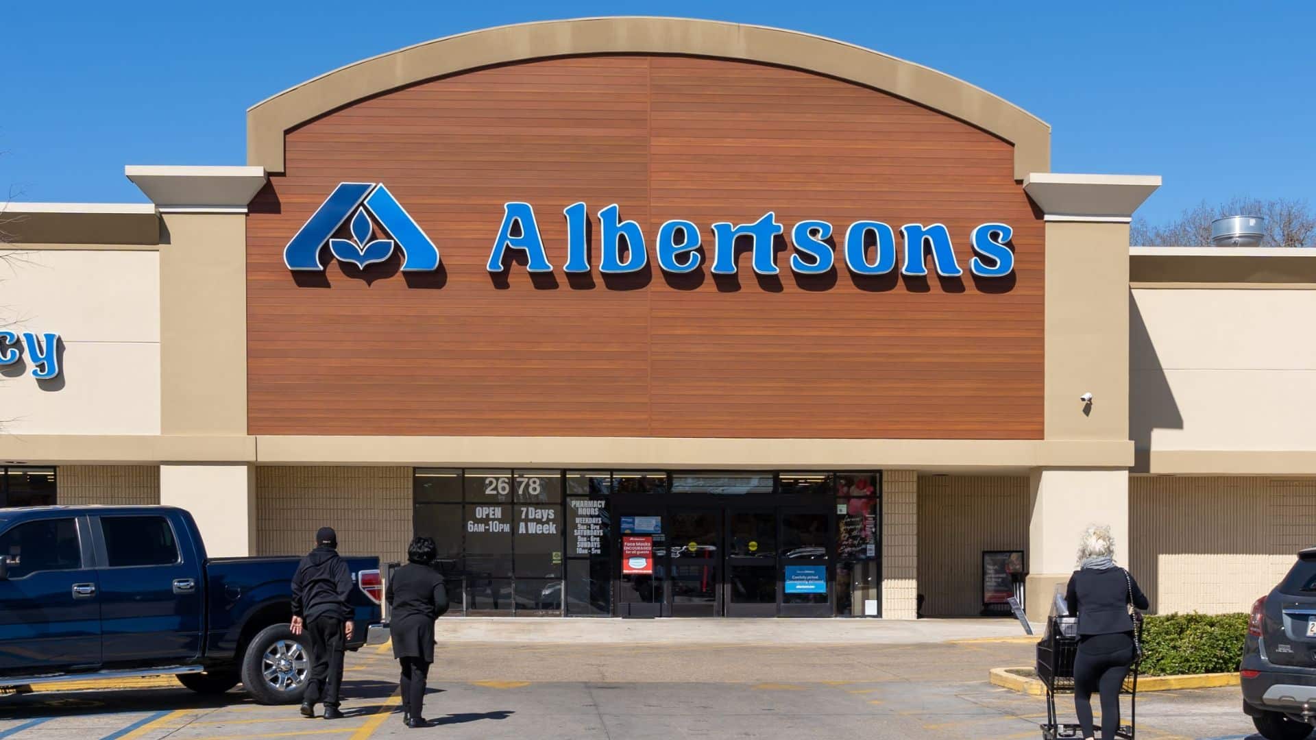 Exterior of an Albertsons store with a large logo above the entrance. People approach the entrance under a clear blue sky.