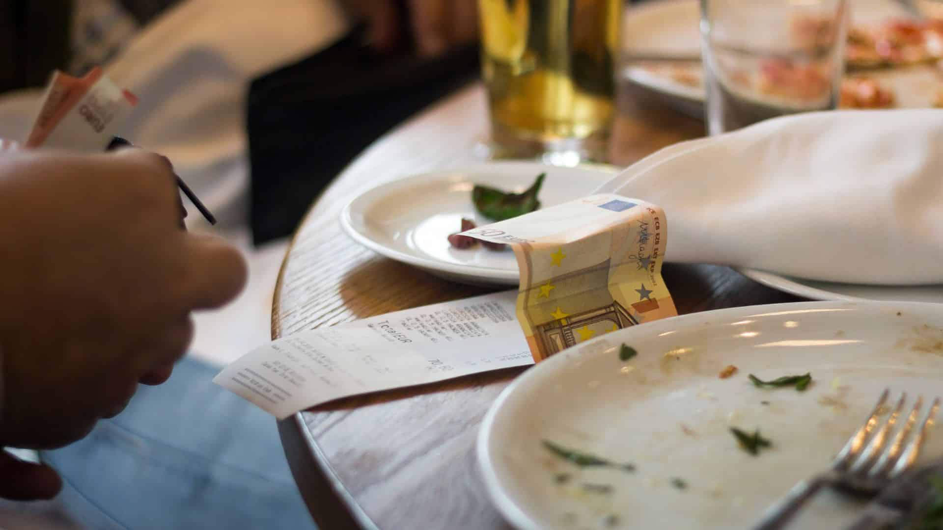 A receipt and banknote on a restaurant table is visible while a diner pulls out money from their wallet.