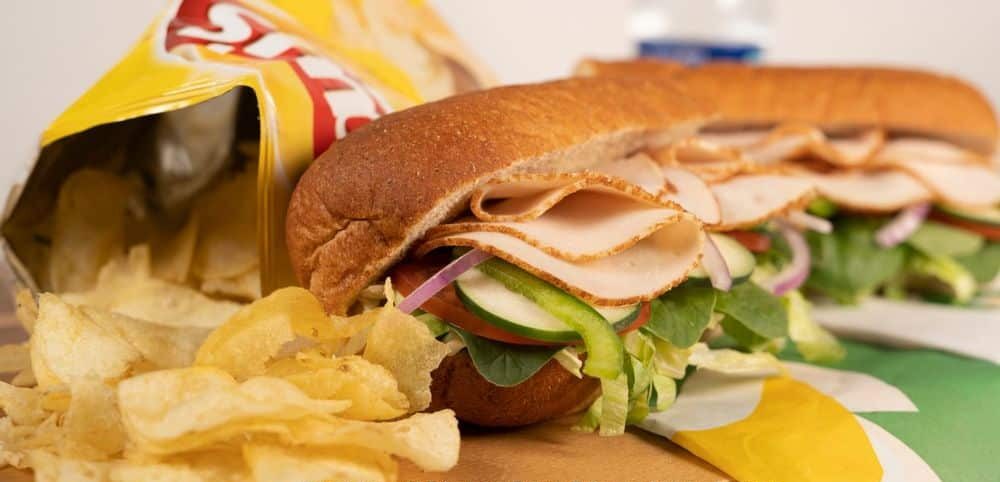 Subway sandwich filled with sliced turkey, lettuce, cucumber, tomato, and red onion on a soft roll, served with a bag of potato chips spilling onto the table beside it.