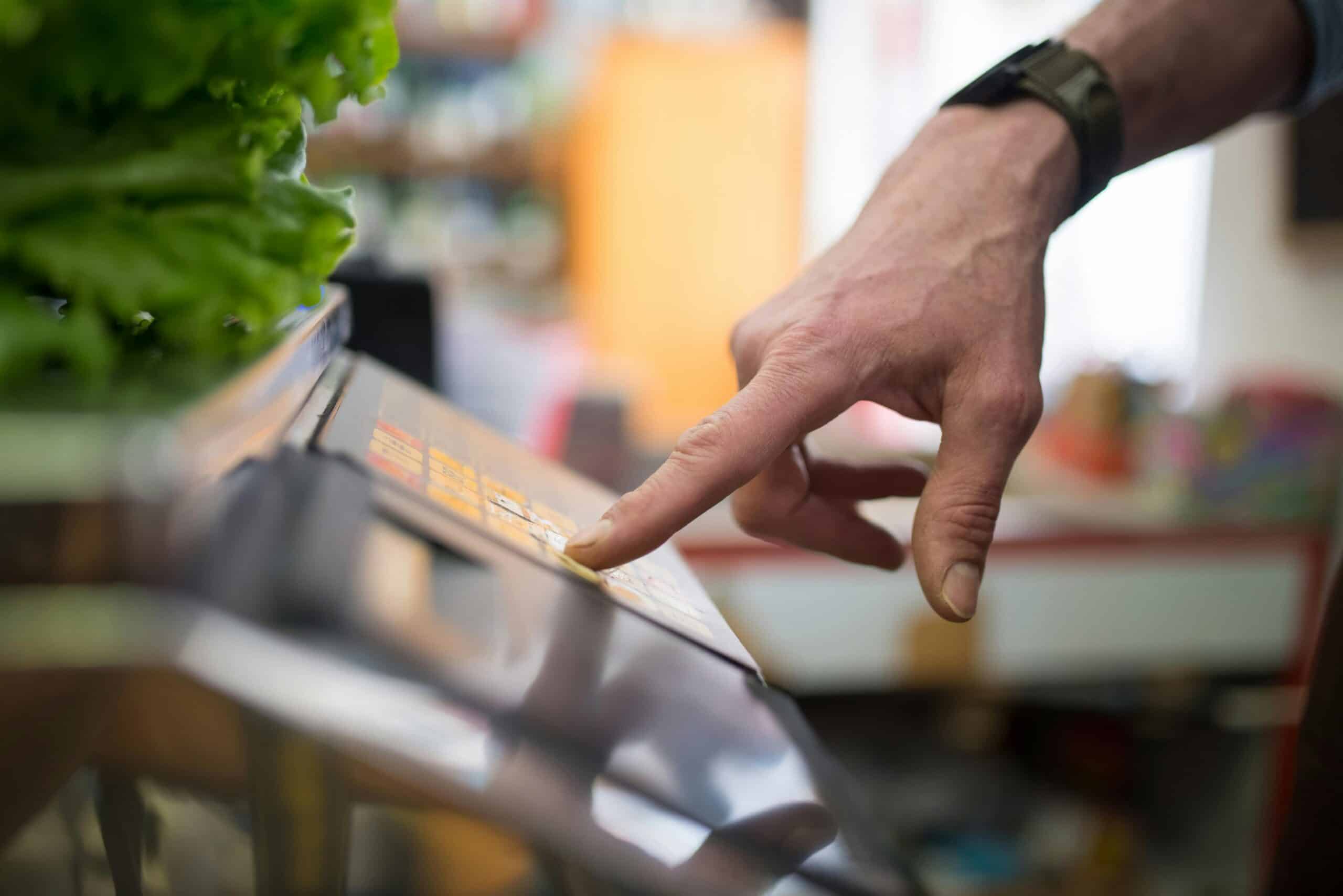 Close-up of a shopper using a touchscreen price-check or self-service display inside a grocery store produce section.